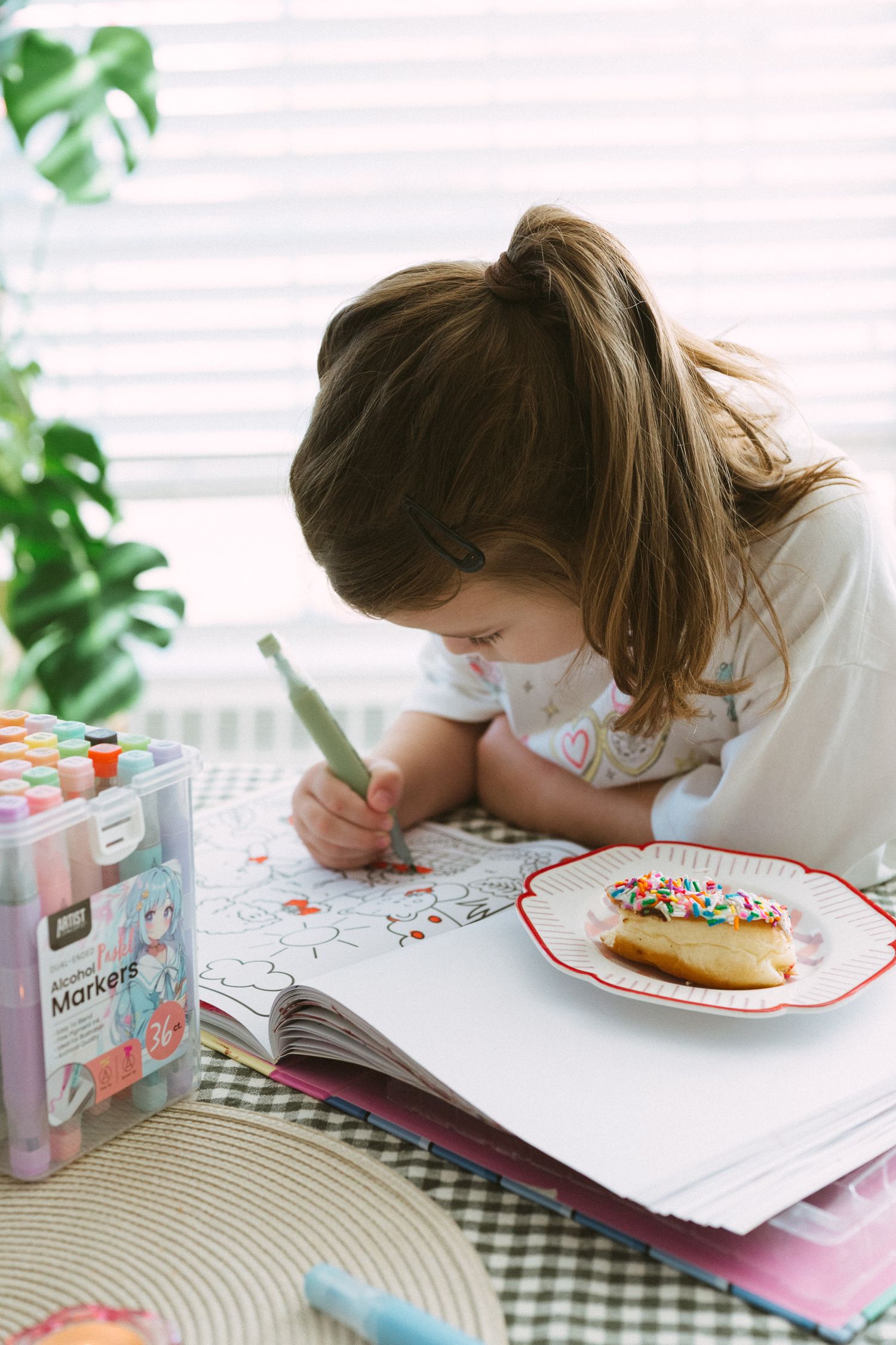 Child lying on table reading or writing in notebook with colorful plate and tropical plants in background.