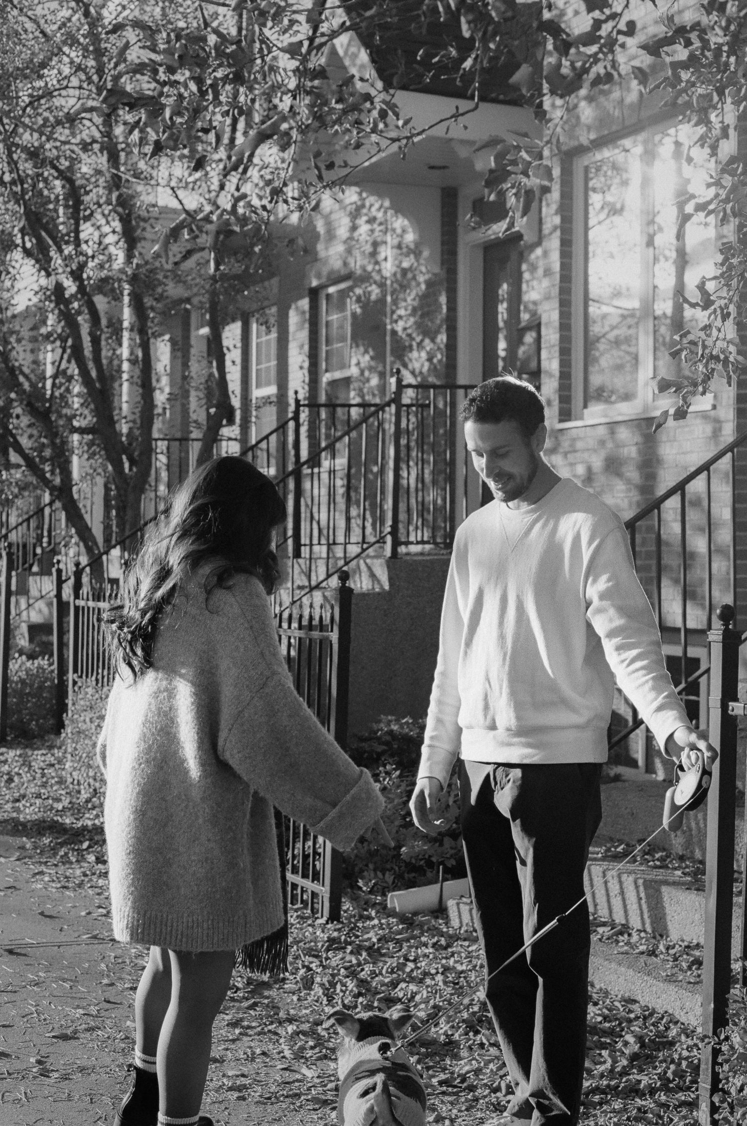 Two people raking fallen leaves together in a yard during autumn with buildings visible behind them.