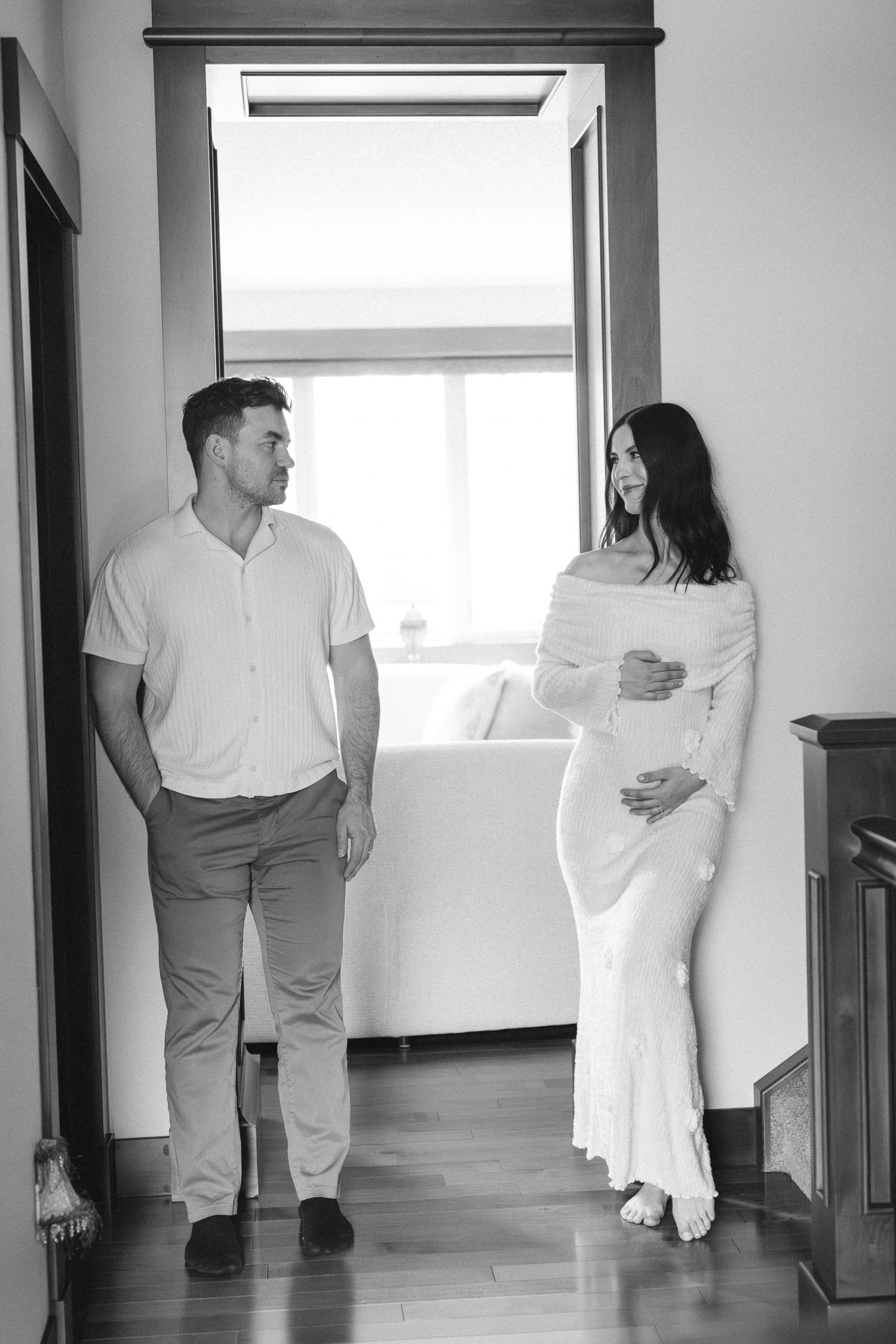 Black and white photo of a pregnant woman in white dress with partner standing by window indoors.