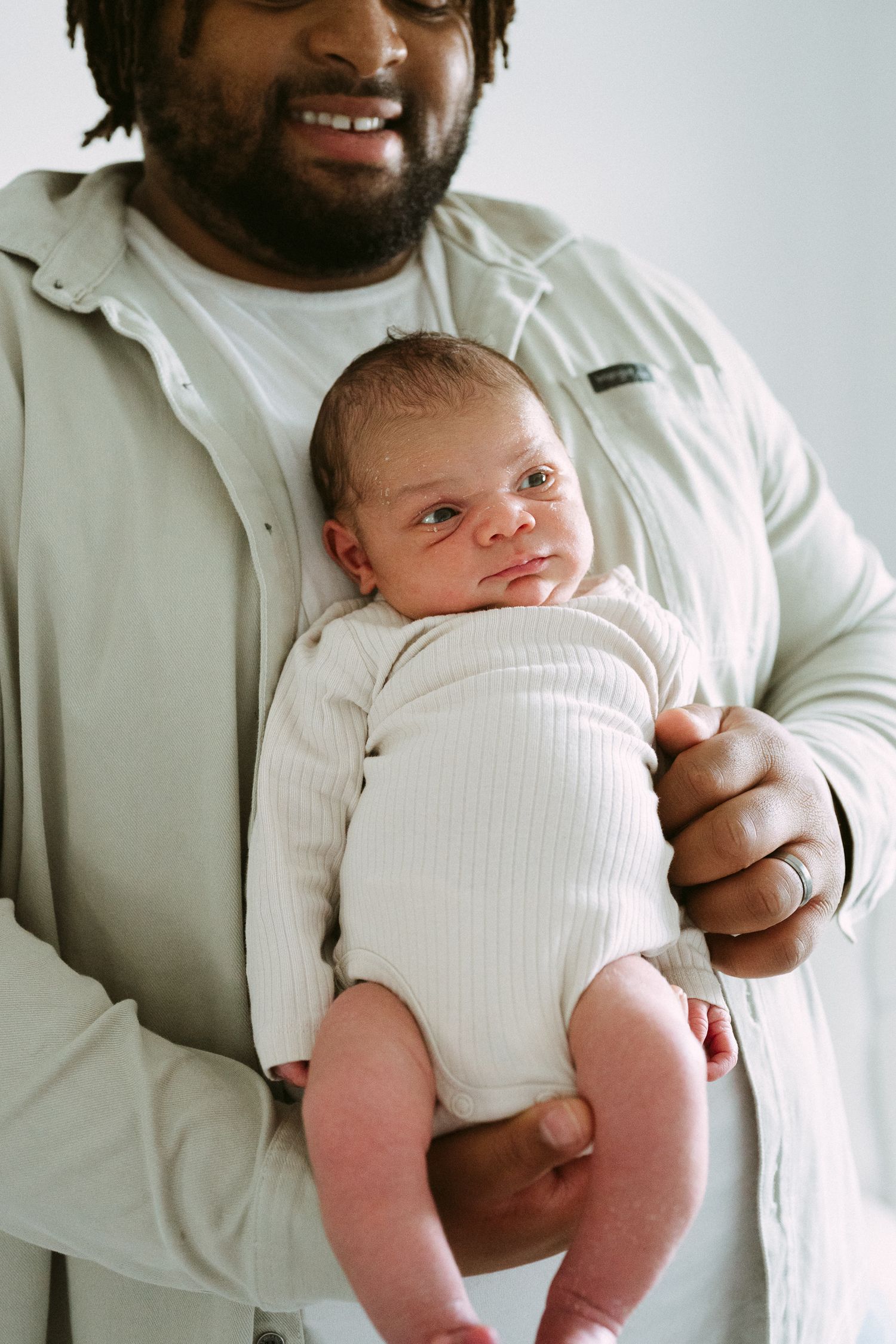 Smiling Black father holding newborn baby dressed in white ribbed onesie against light background.