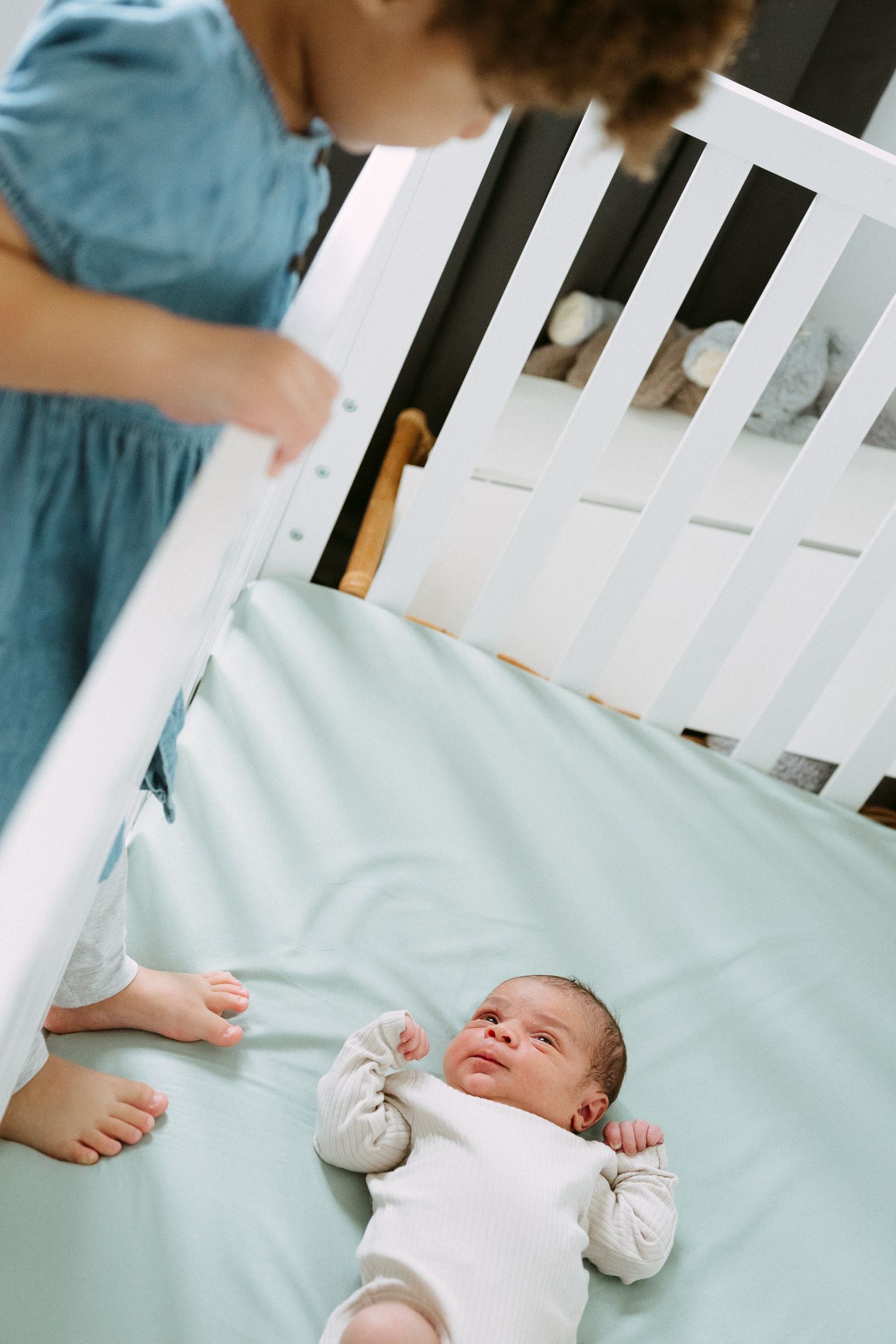 Mother in blue dress leaning over white crib with newborn baby lying on mint green sheet.