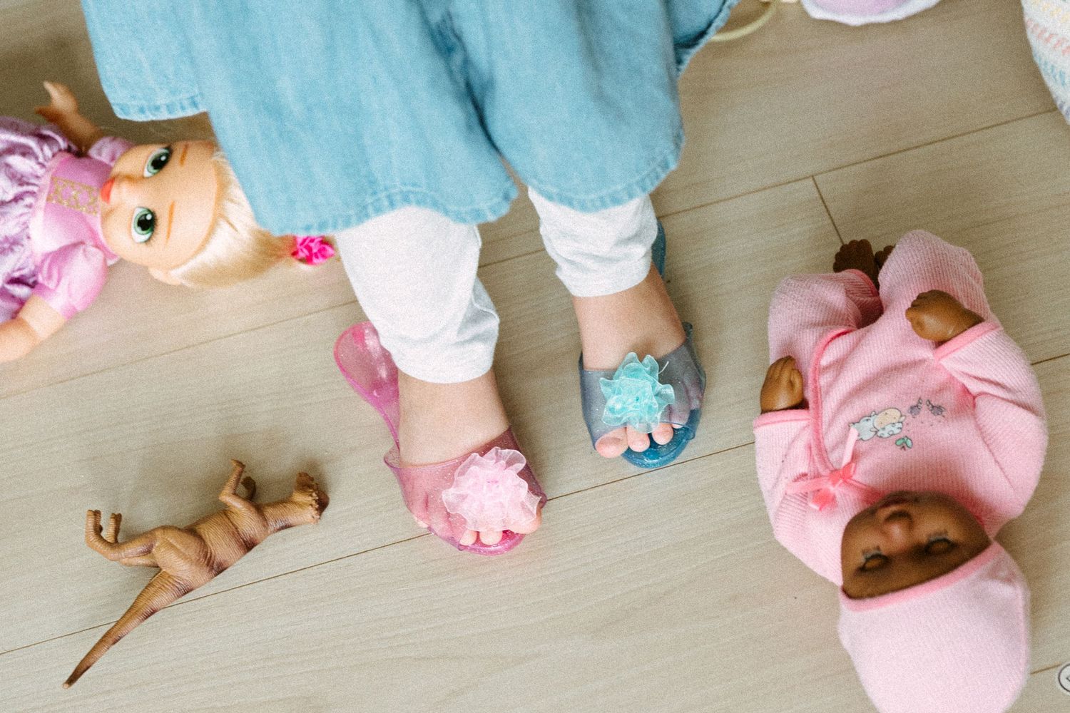 Child wearing colorful slippers standing among scattered baby dolls on a light wooden floor.