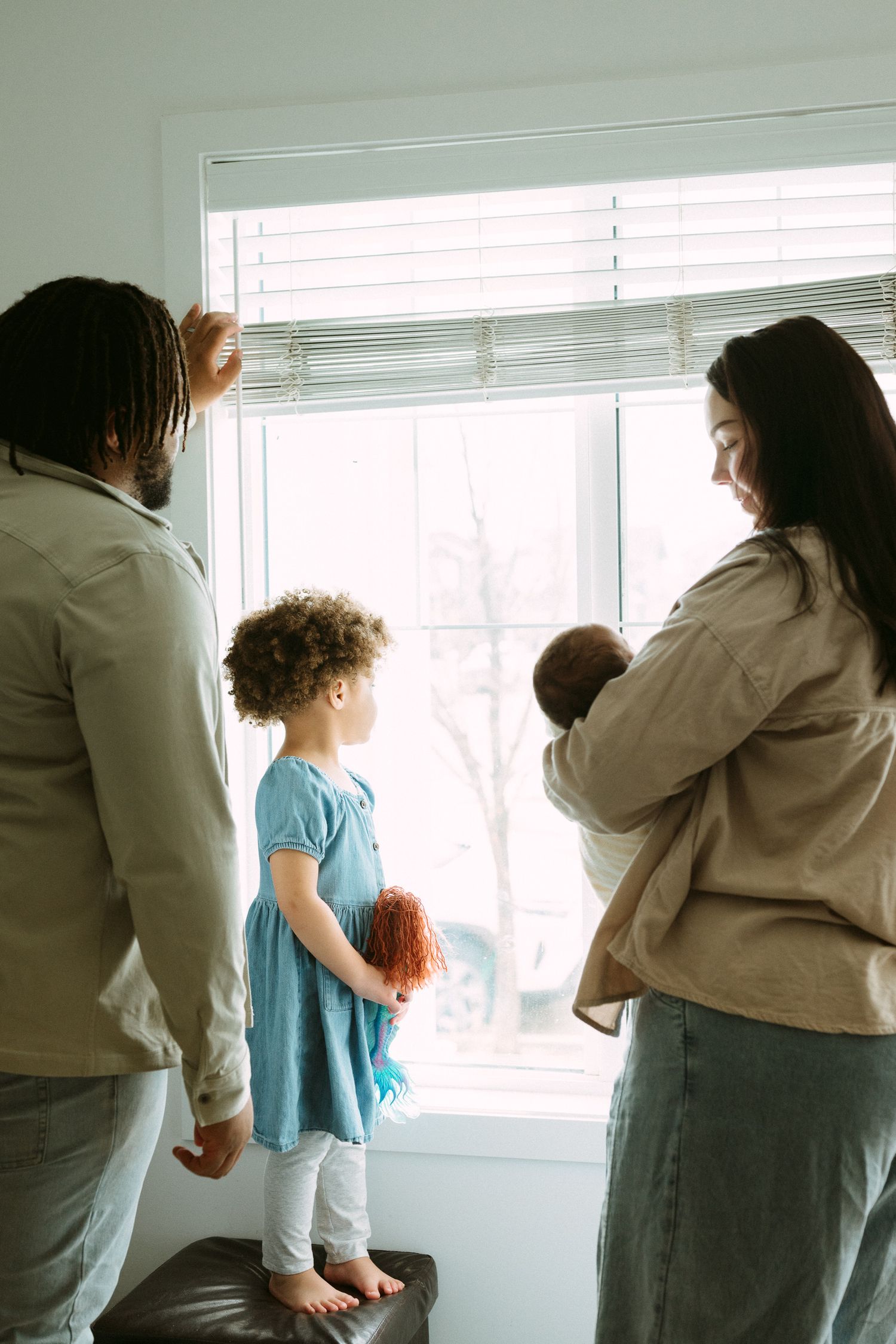 A family of four looks out a bright window together, father holding daughters hand, mother cradling newborn.
