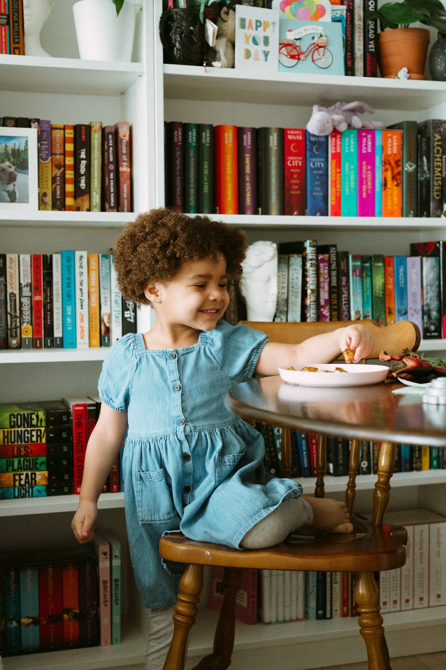 A young girl with curly hair in a blue dress sits at a wooden table eating, with colorful bookshelves behind her.