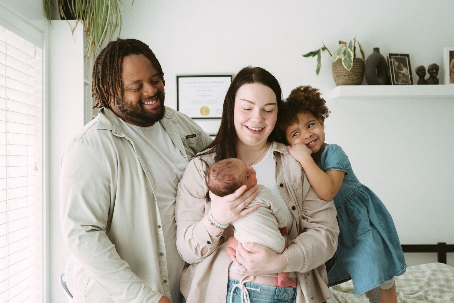 A joyful family of four poses indoors, parents smiling with a newborn and young daughter in a blue dress.