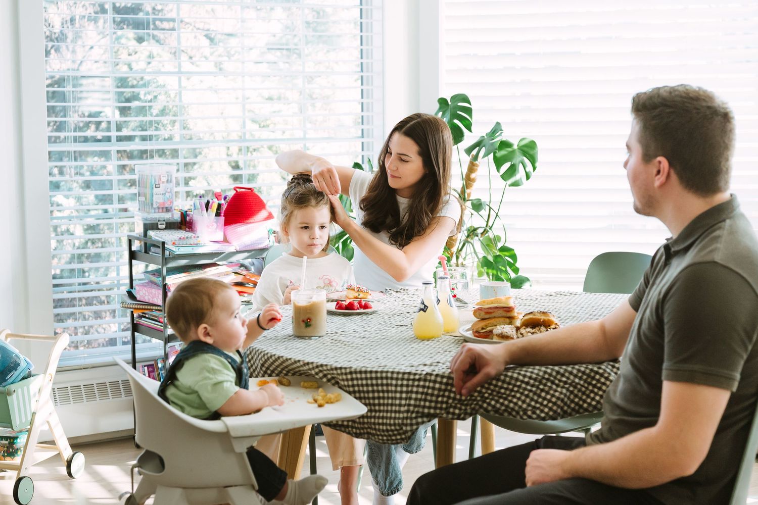 Family gathered around dining table in bright sunlit room with tropical plants and windows.