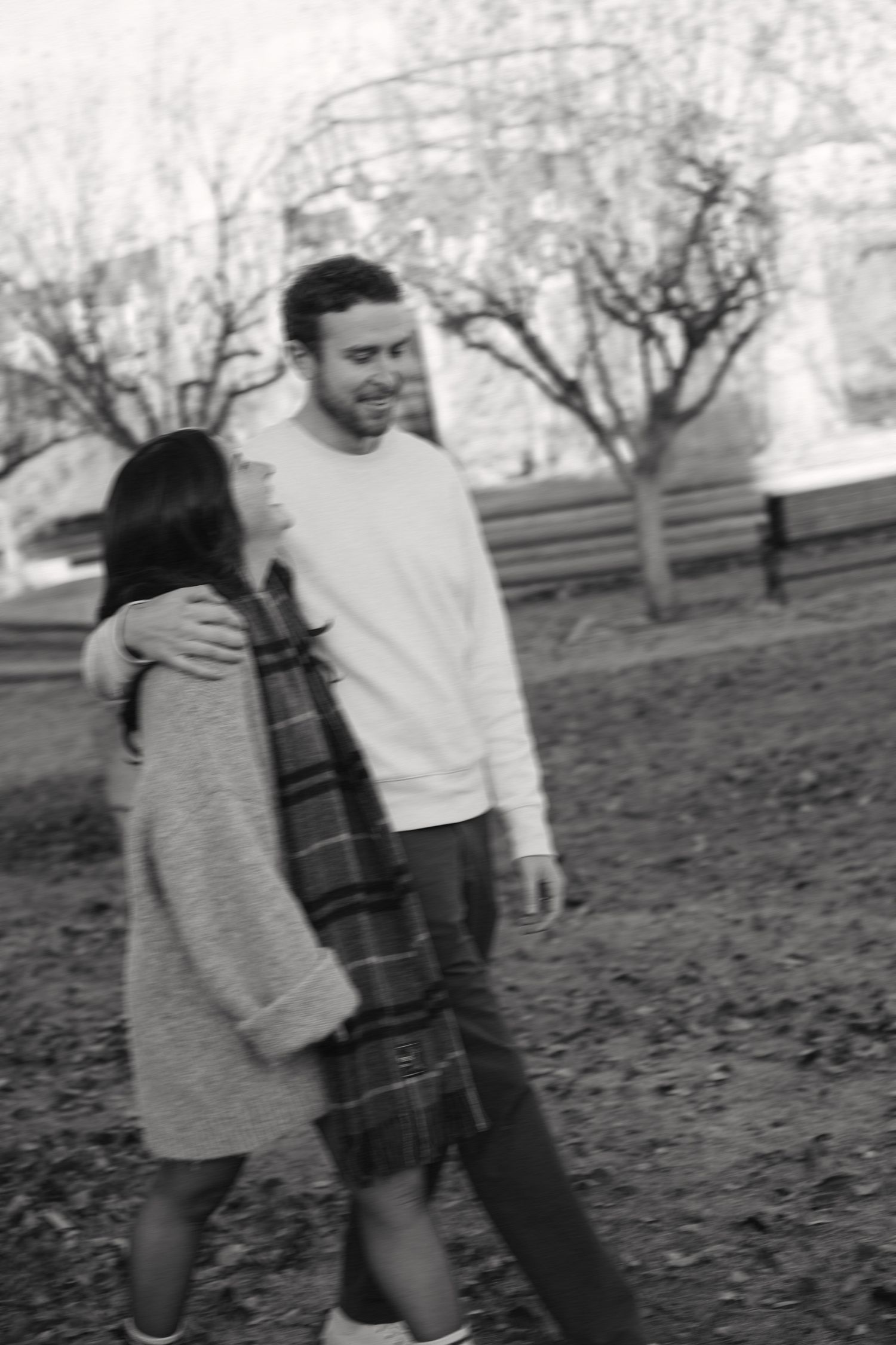 A couple embraces while standing in a park during autumn with bare trees visible in the background.