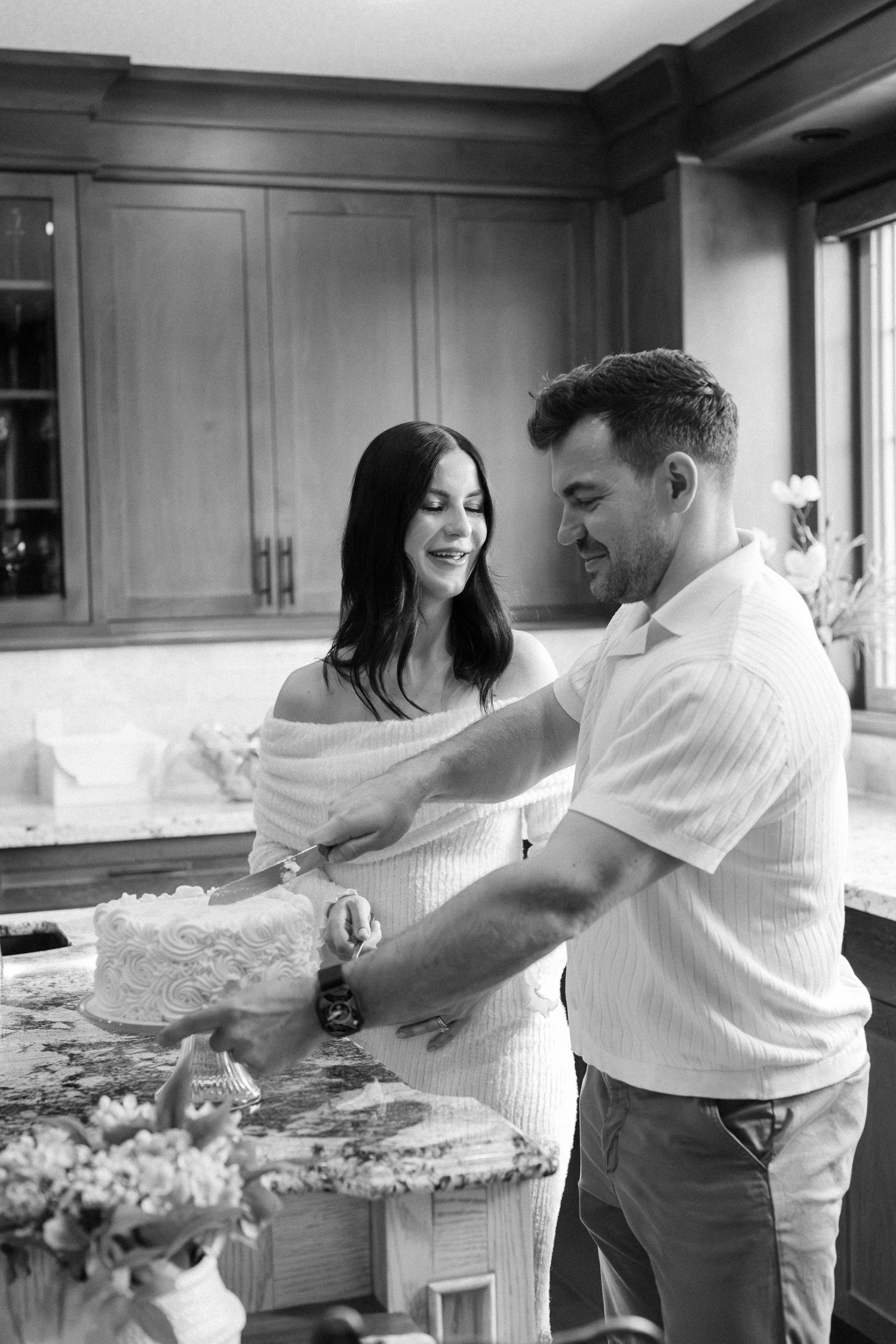 Black and white photo of a couple cutting a wedding cake together in an elegant kitchen setting.