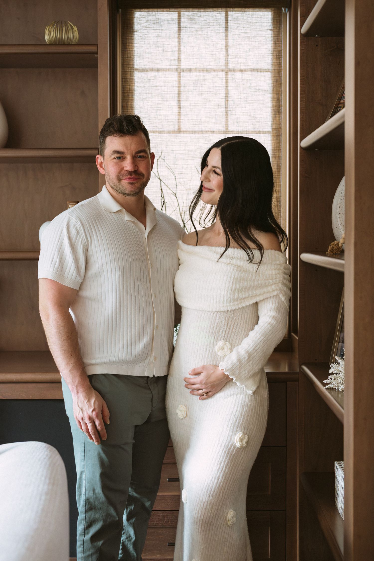 Couple posing for maternity photos in a cozy wood-paneled room, woman in white off-shoulder dress.
