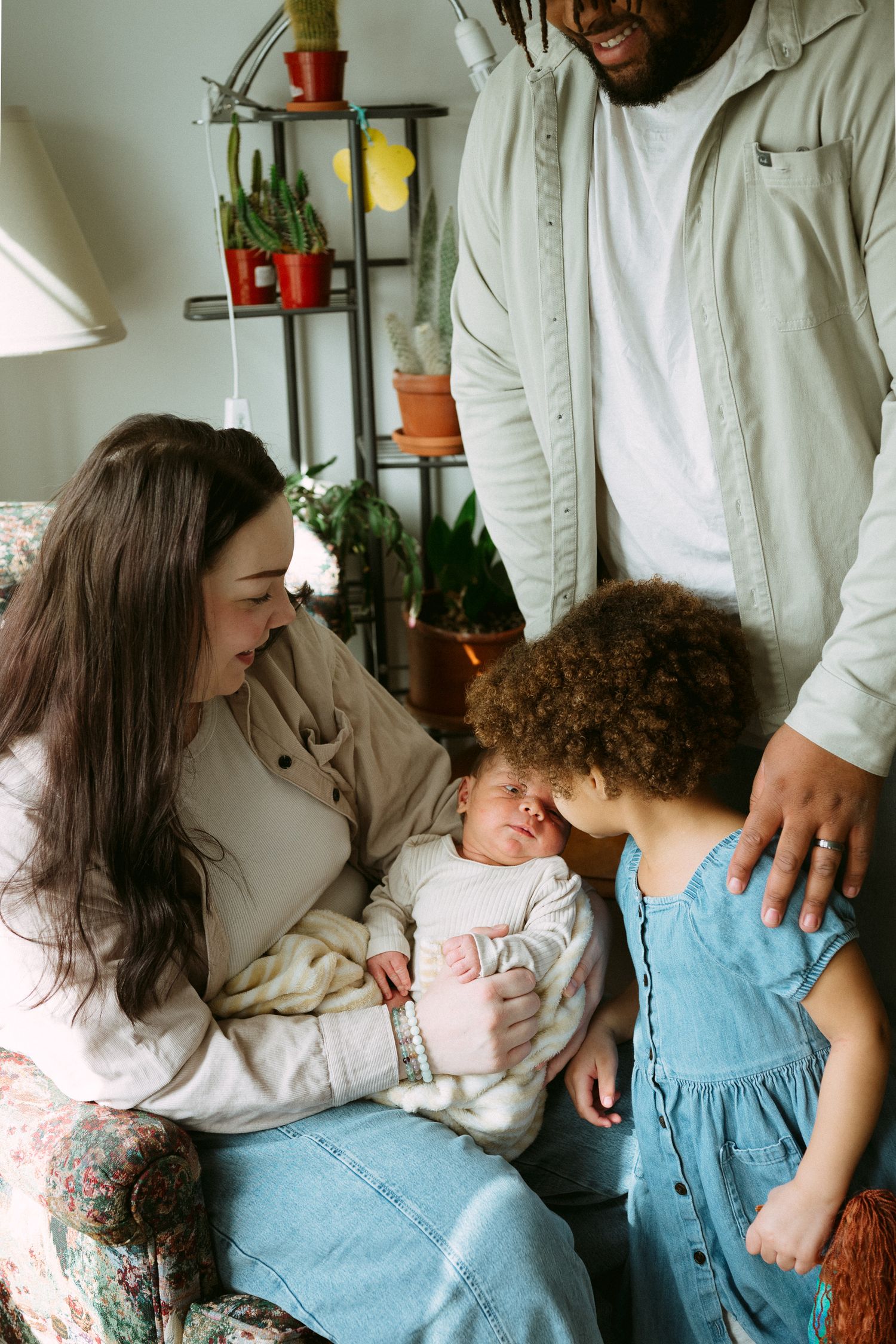 A young family admires their newborn baby at home, with mother seated holding infant while father and child look on lovingly.