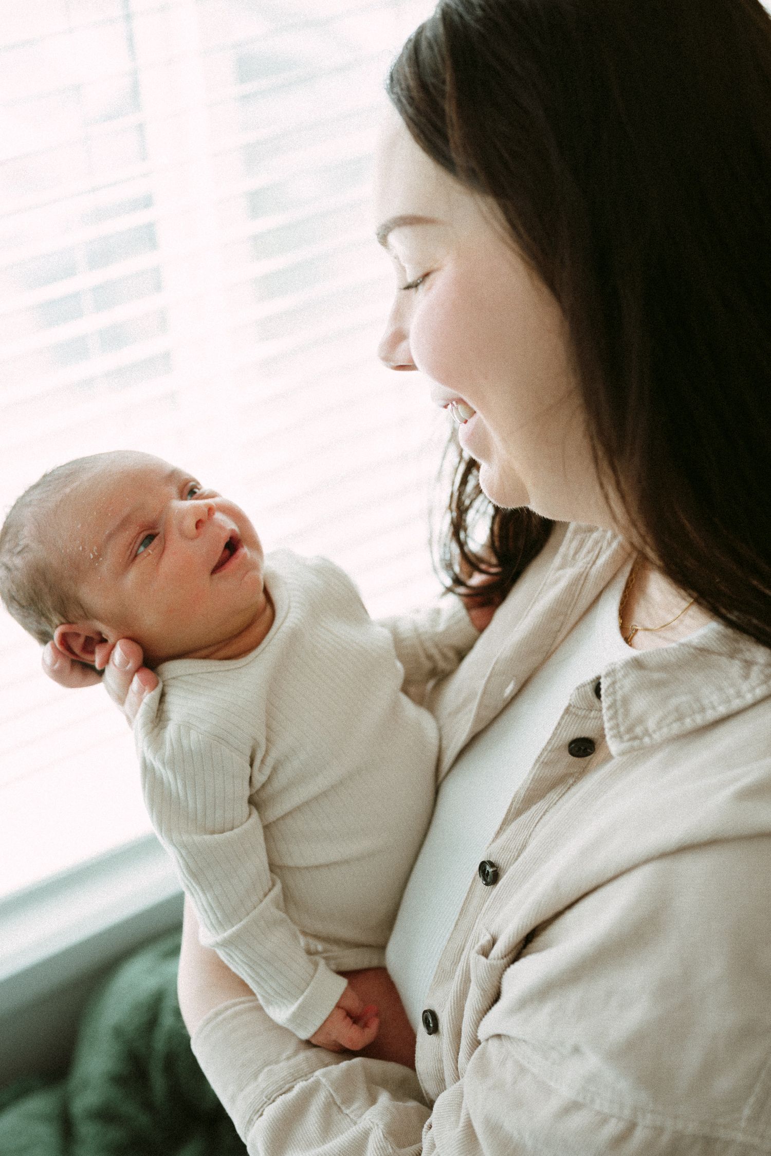 Smiling dark-haired mother holding and gazing at her newborn baby near a bright window.
