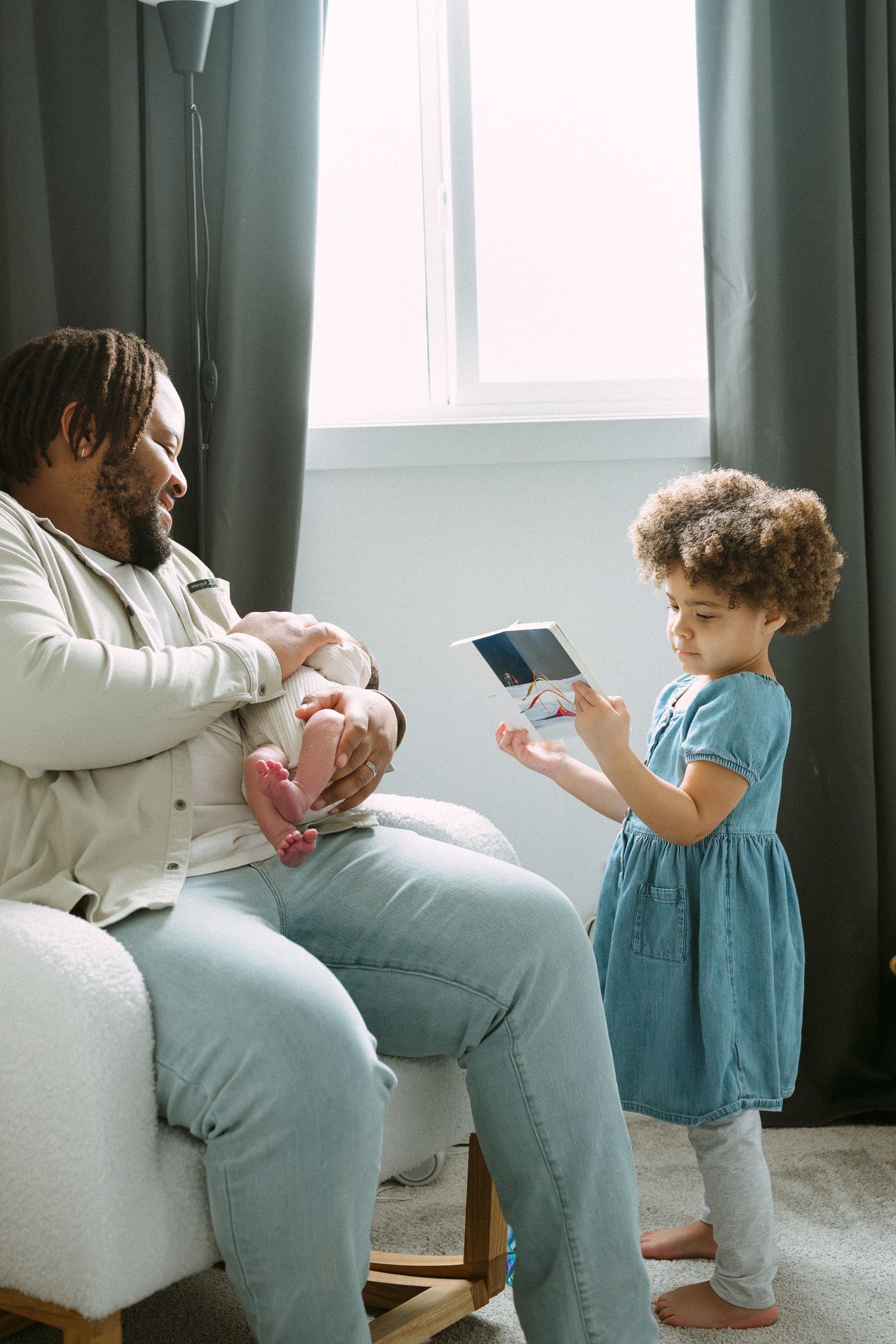 Father sits in nursery chair holding newborn as young curly-haired girl in blue dress shows a book.