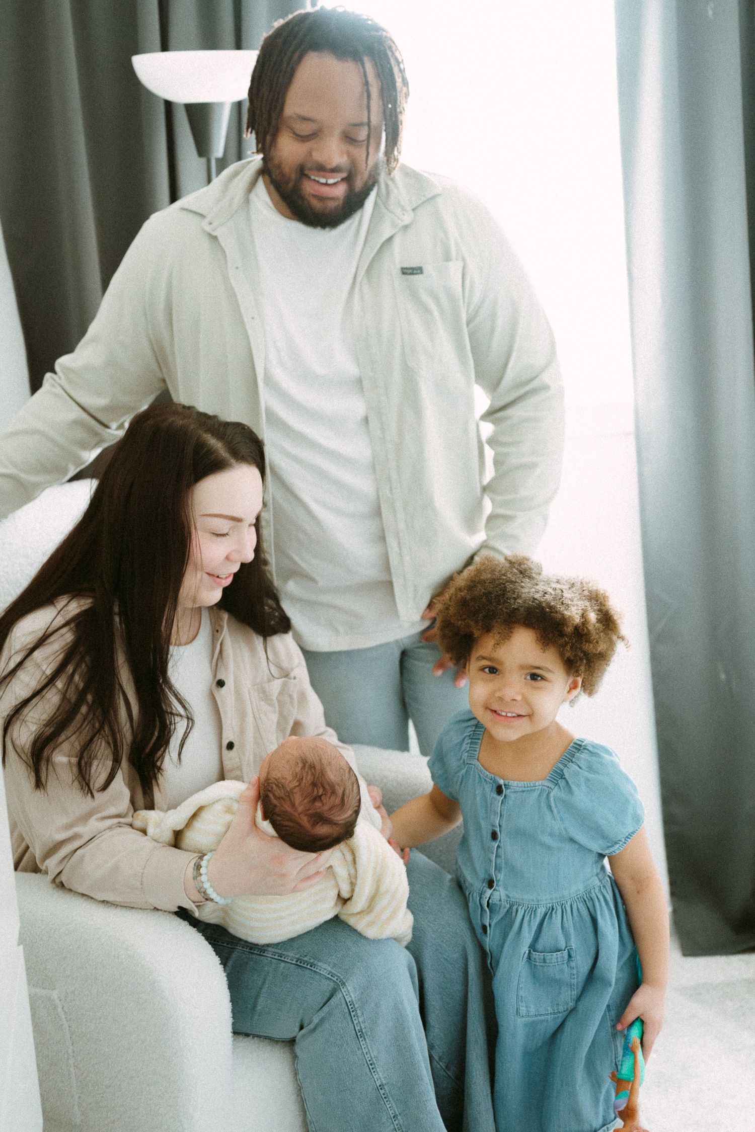 A happy multiracial family admires their newborn baby, with mom seated, dad standing, and young daughter smiling nearby.
