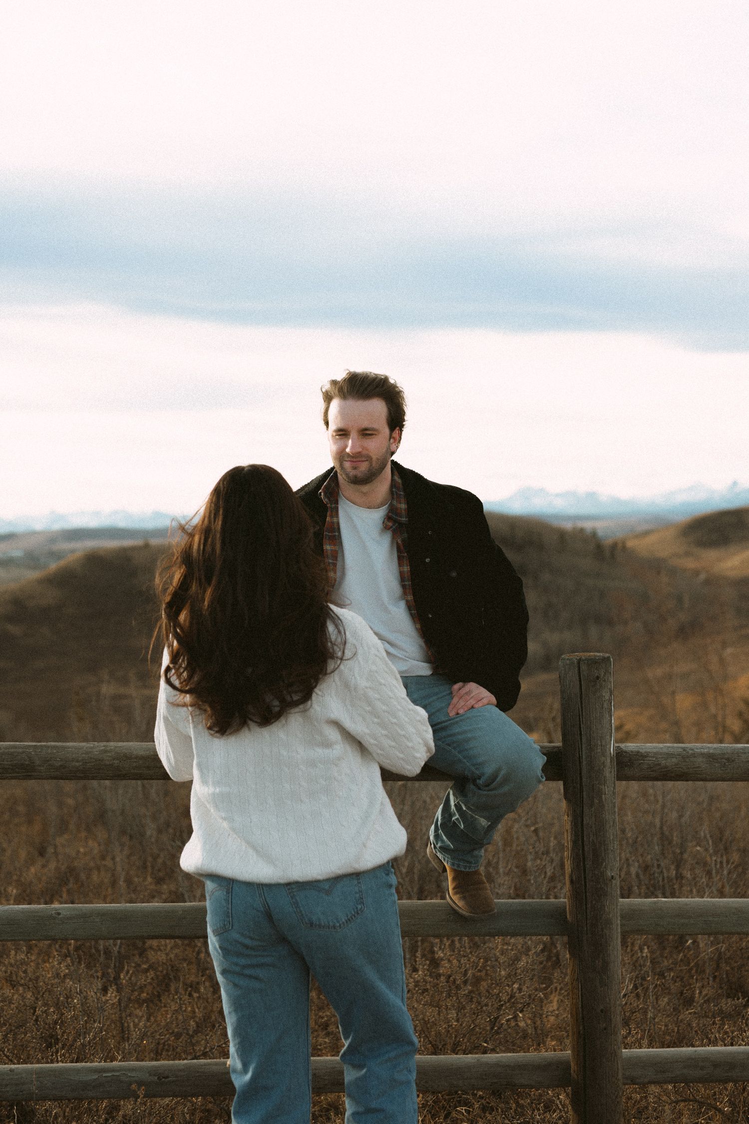 Two people share an intimate moment while sitting on a rustic fence with rolling hills in the background.