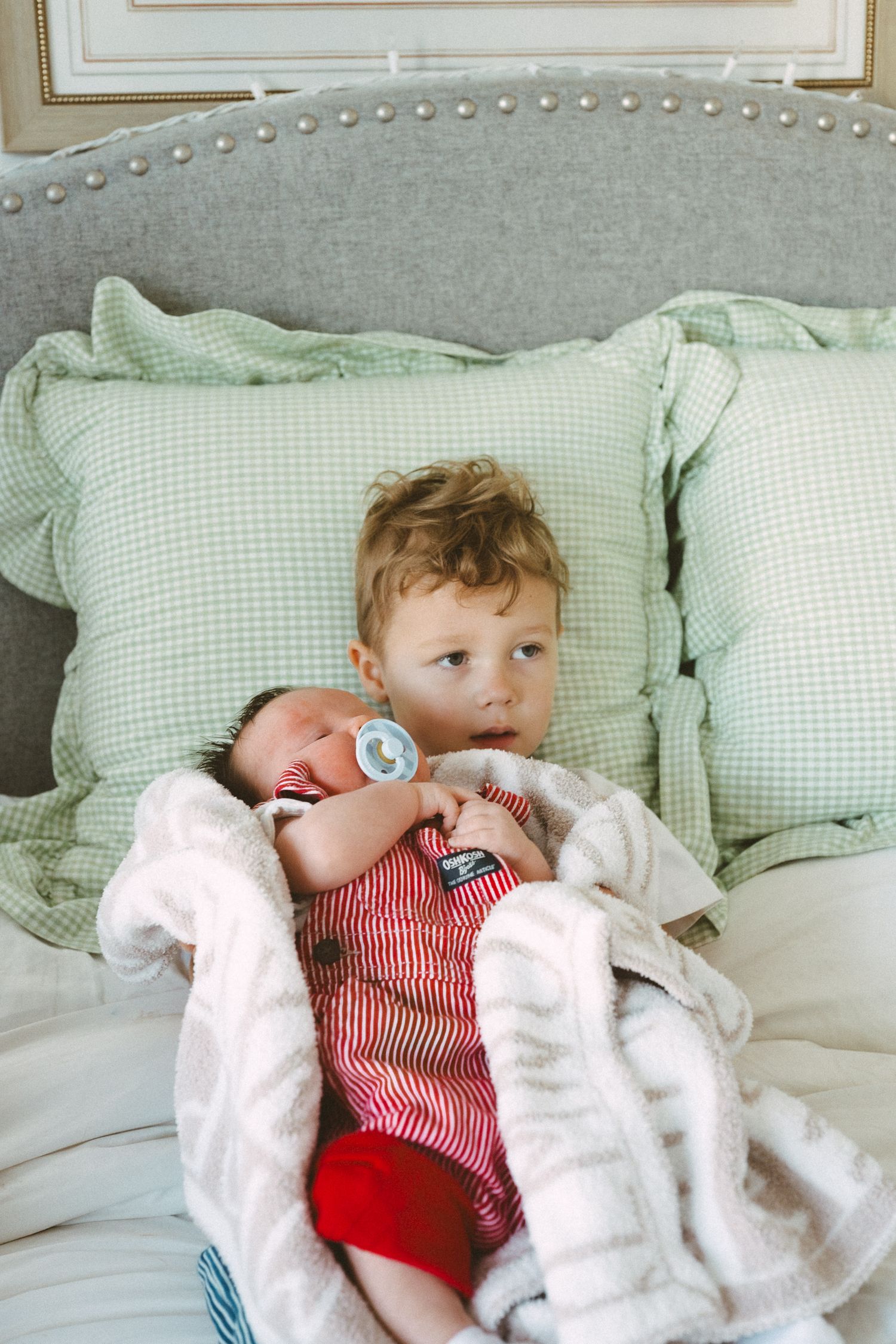 A toddler in red and white striped clothing rests on mint green pillows against an upholstered headboard.