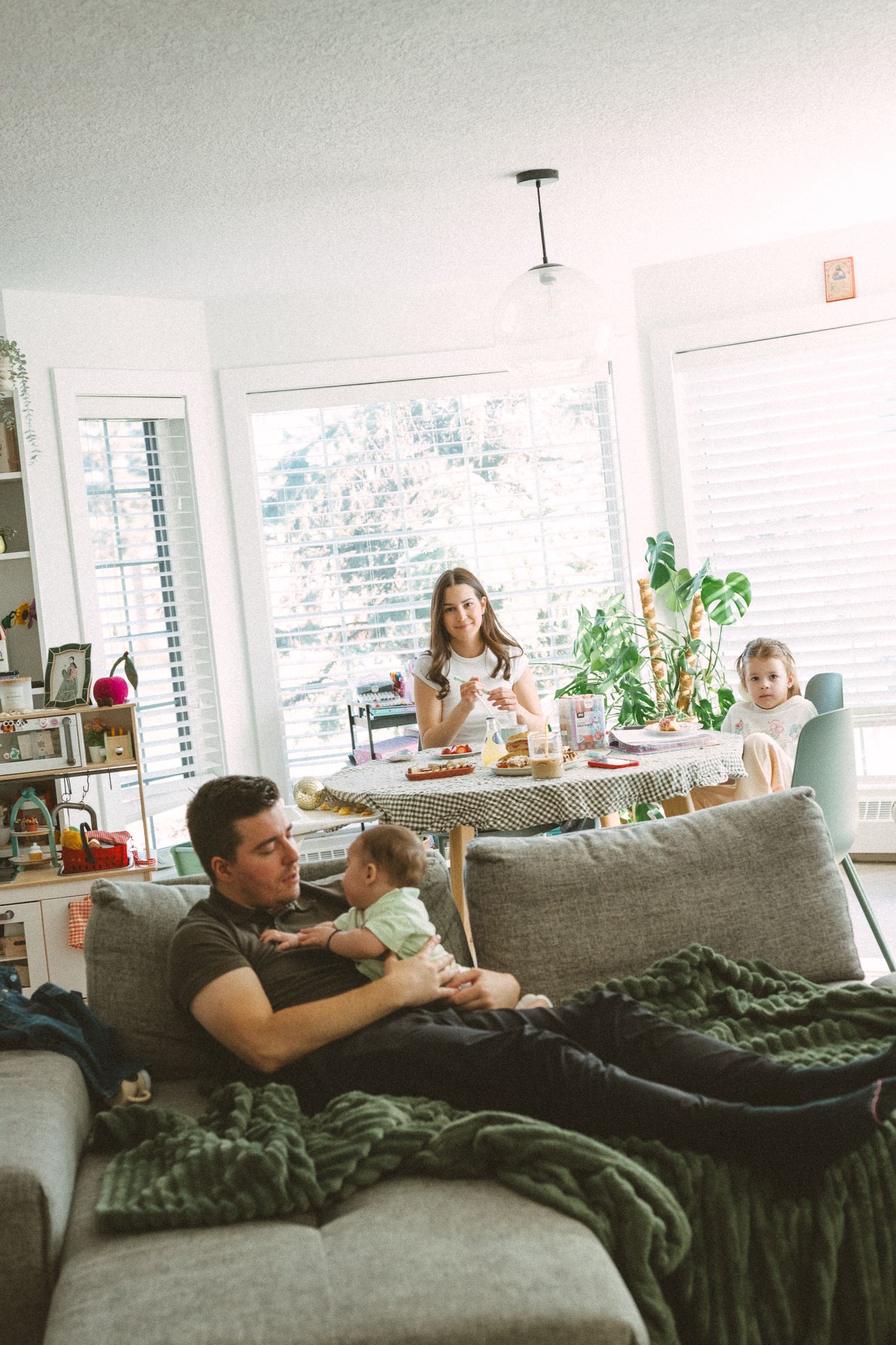 Family relaxing together on a grey couch with a green knit blanket in a bright sunlit living room.