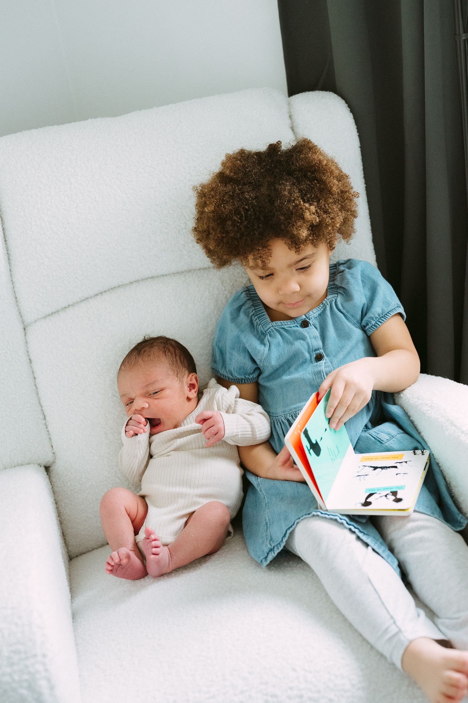 A young curly-haired girl in a denim dress reads a colorful board book to a newborn baby on a white chair.