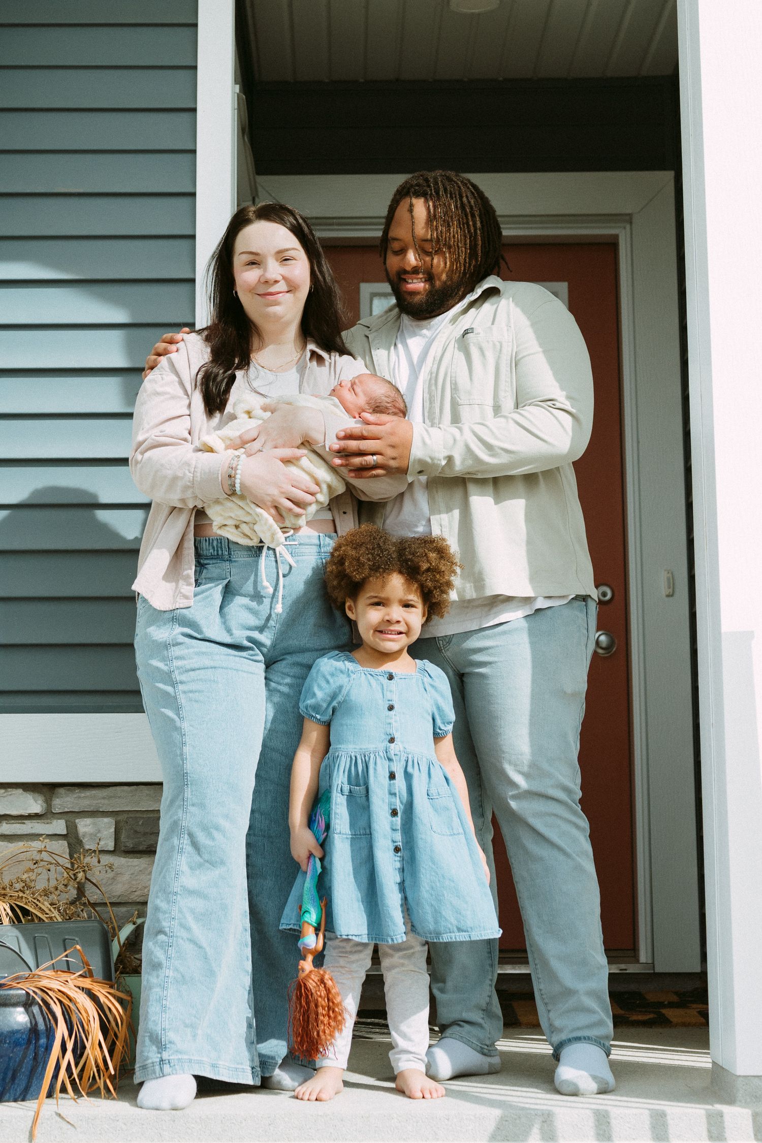 Interracial family of four posing on front porch, parents holding newborn baby with toddler daughter standing in front.