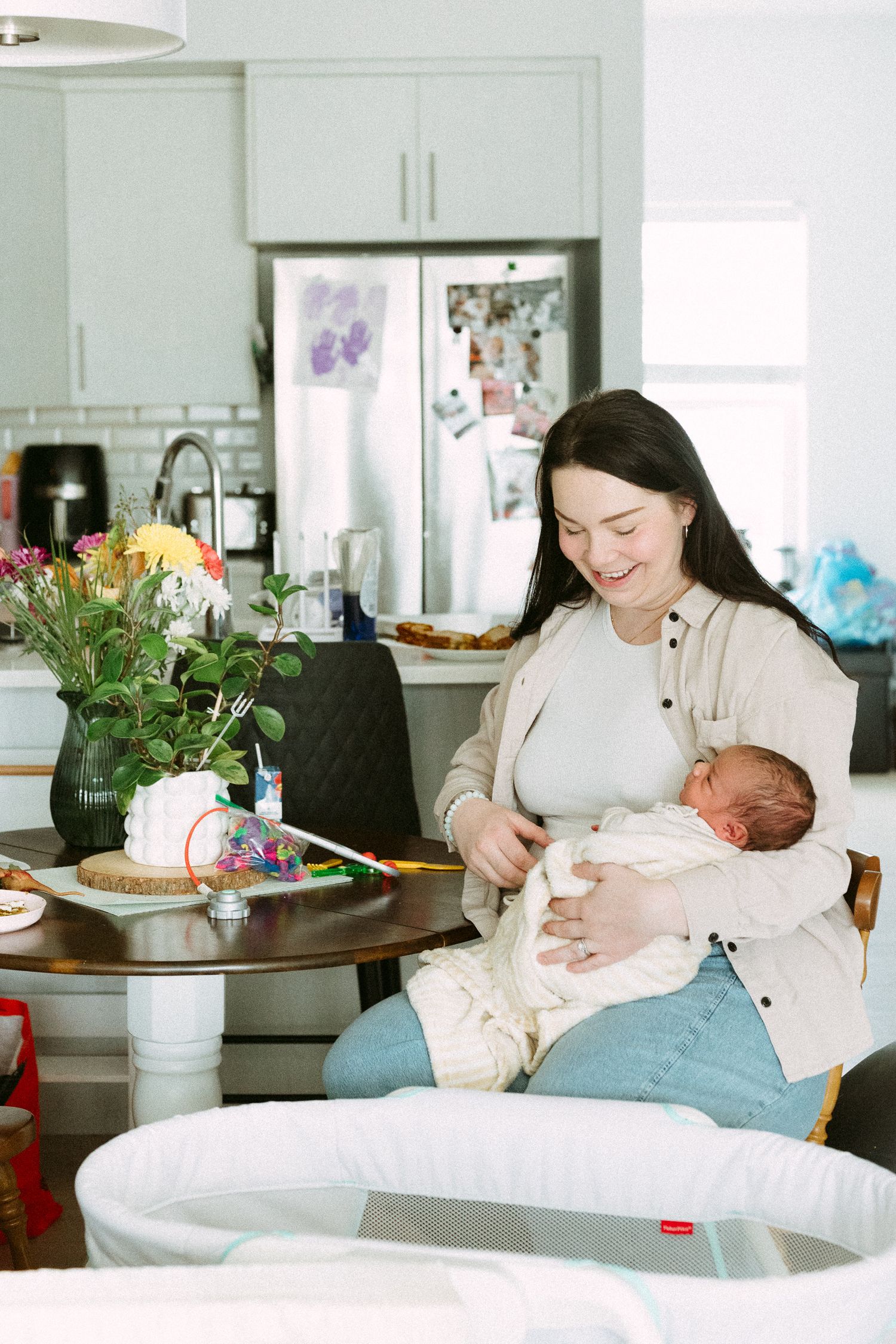 A smiling woman with dark hair holds her newborn baby while sitting at a kitchen table with flowers nearby.