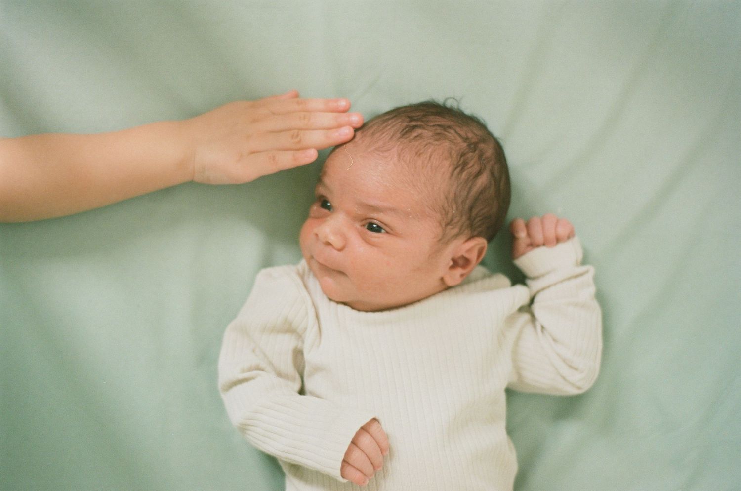 Newborn baby in white onesie lying on green blanket while parent gently touches their head.