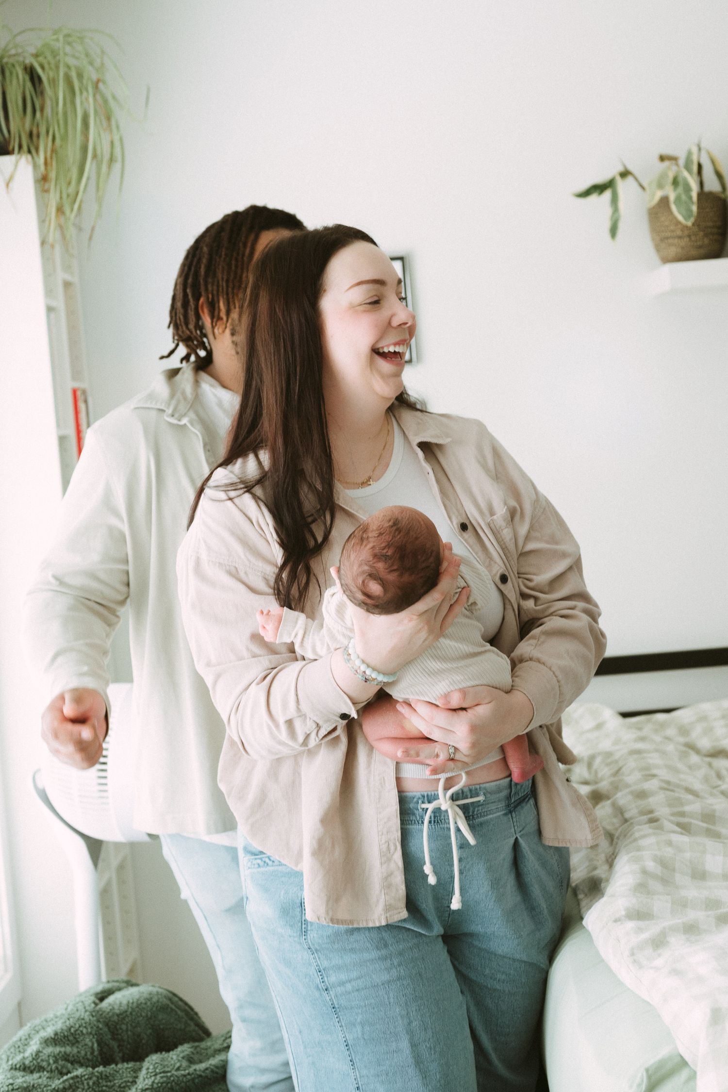 A laughing mother holds her newborn while her partner stands close behind her in a bright, cozy bedroom.