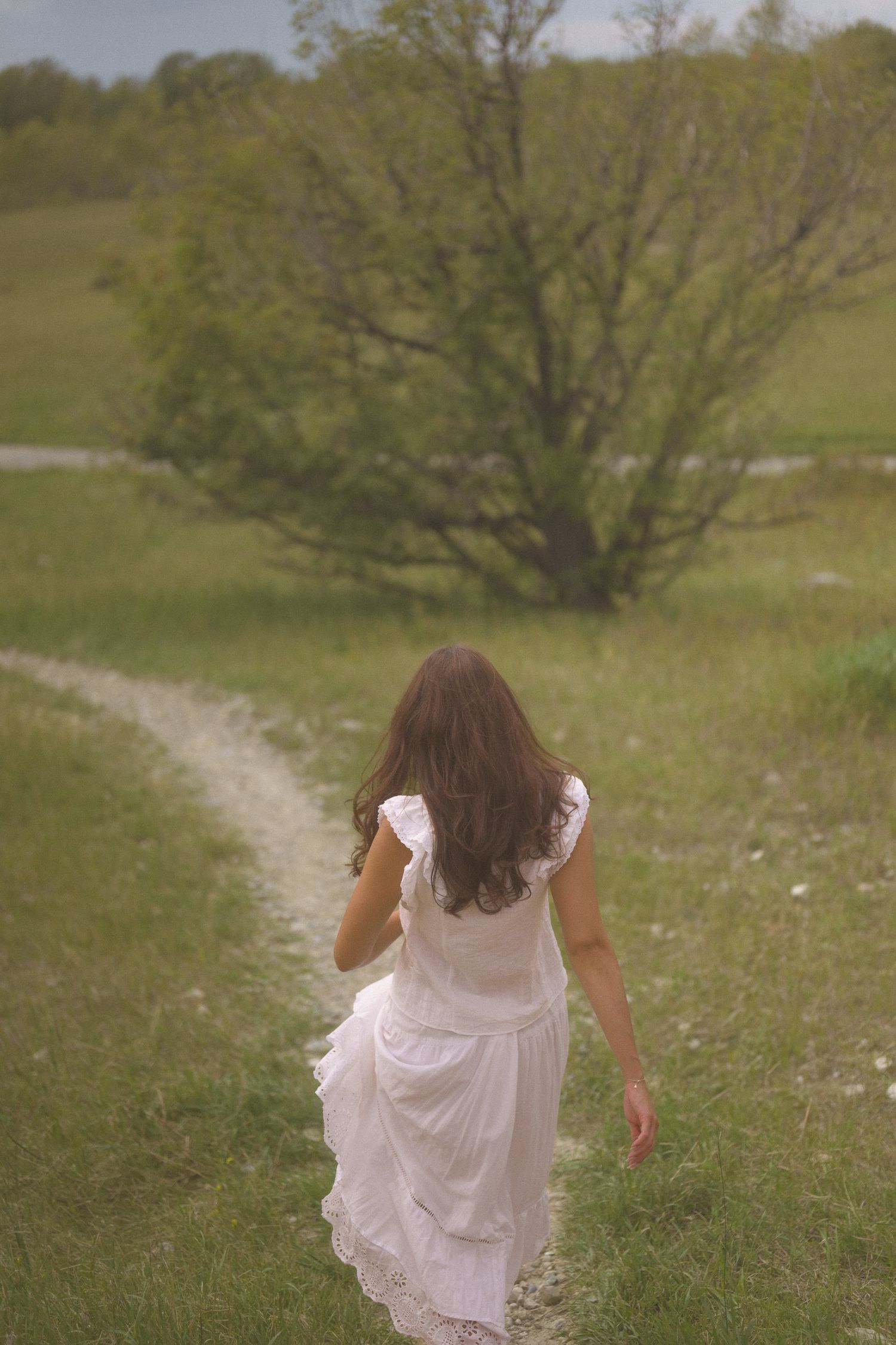 A person in a flowing white dress walks away down a nature path surrounded by green grass.
