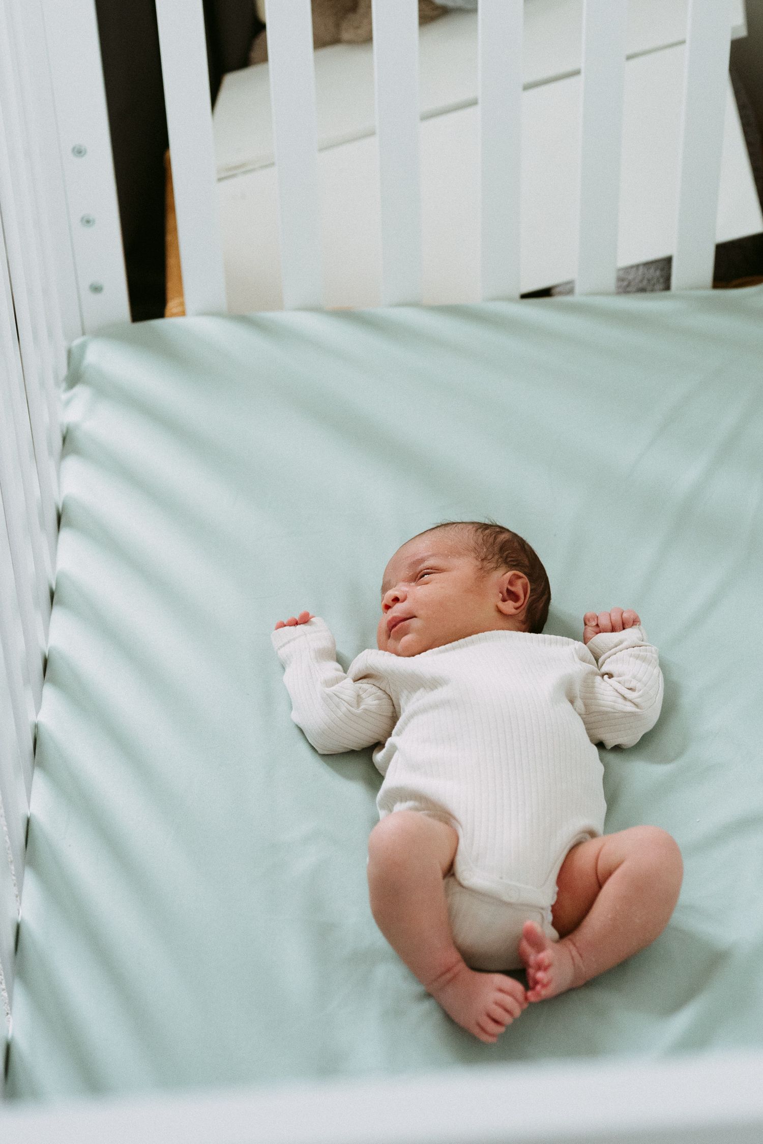 Newborn baby in white onesie lying peacefully in a white crib with mint green sheets.