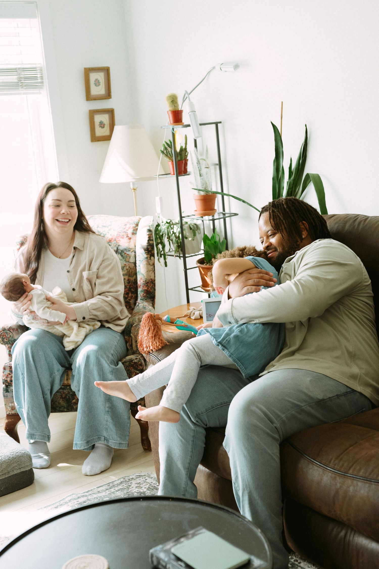A happy interracial family relaxes on sofas in a cozy living room, with parents holding young children and laughing together.