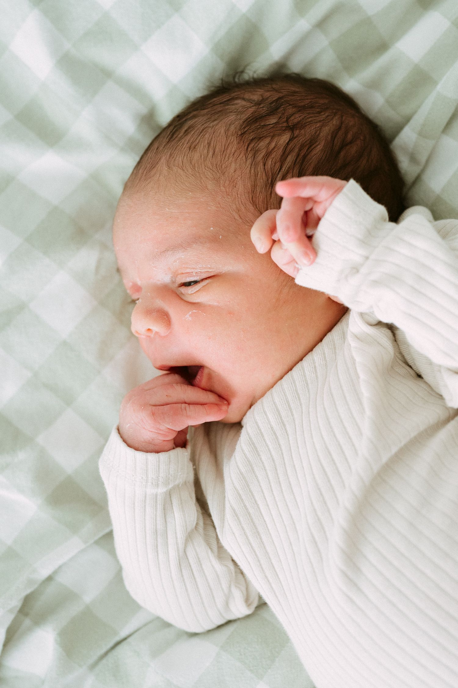 Newborn baby in white ribbed outfit yawning with hand near face, lying on green gingham blanket.