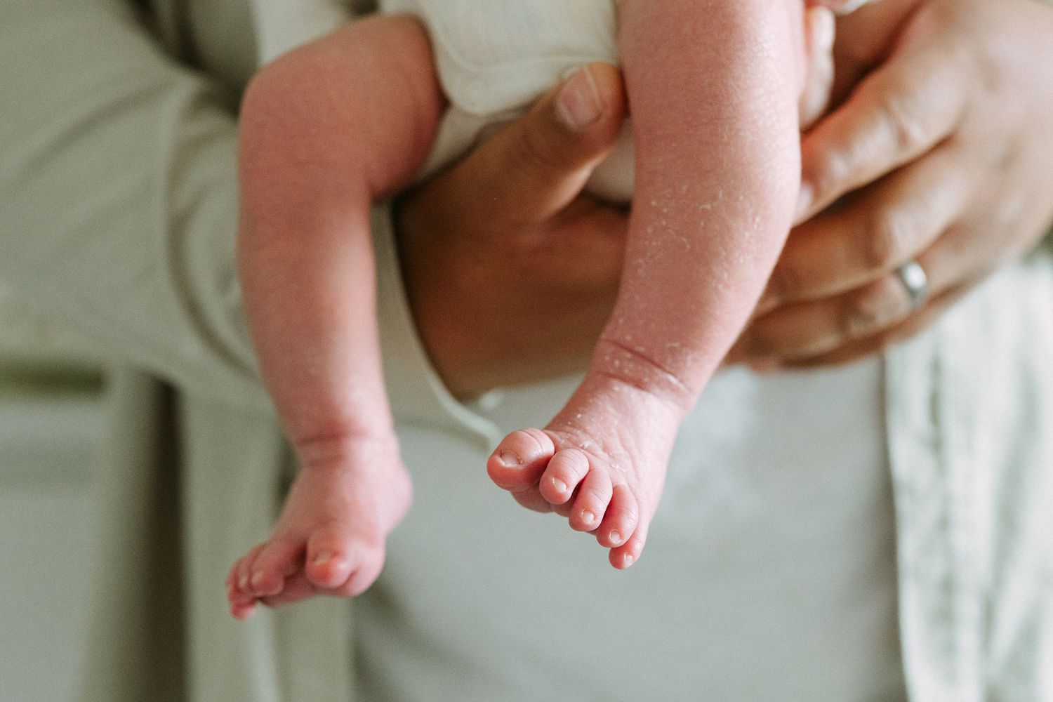 Close-up of newborn baby feet and legs being held securely by a parent wearing a wedding ring.