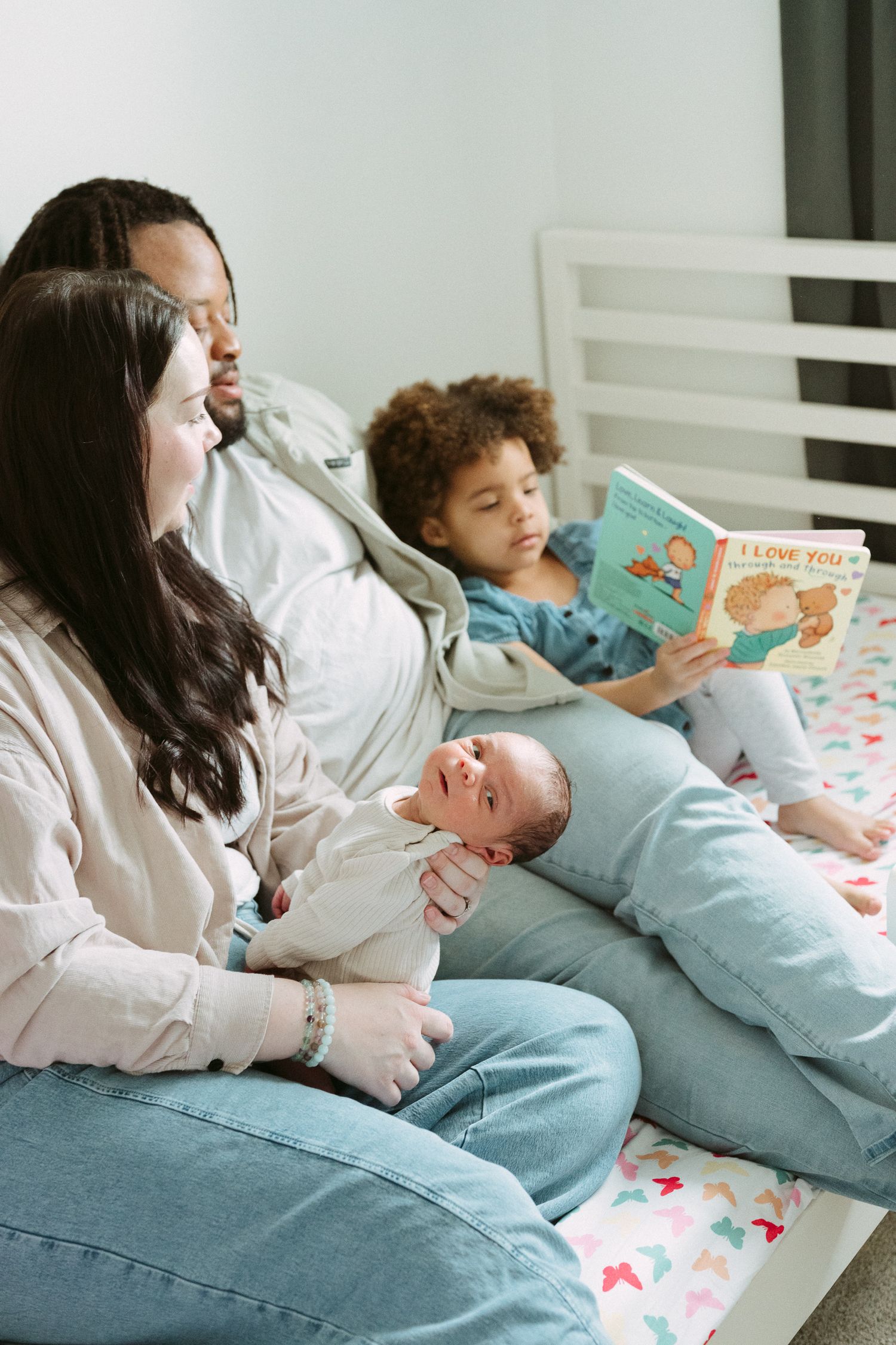 Family sitting on bed with newborn baby while older child reads I Love You picture book together.