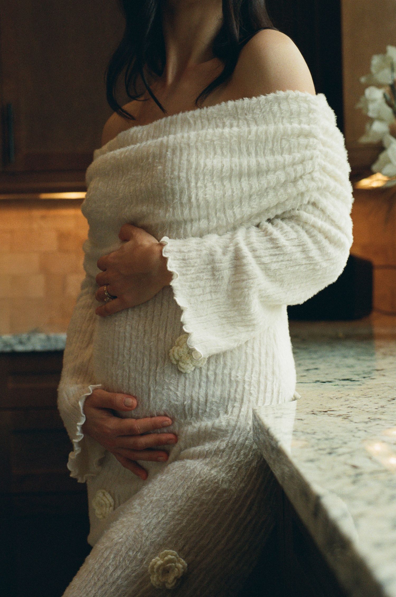 Pregnant woman in off-shoulder cream textured knit dress holding baby bump beside marble kitchen counter.