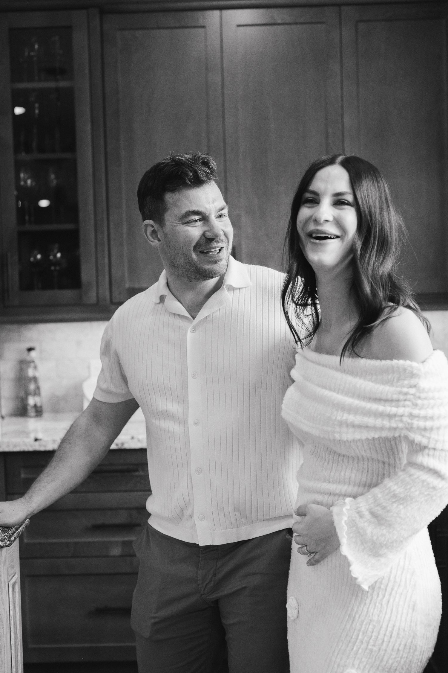Happy couple laughing together in a kitchen, black and white maternity photo with woman in off-shoulder white dress.