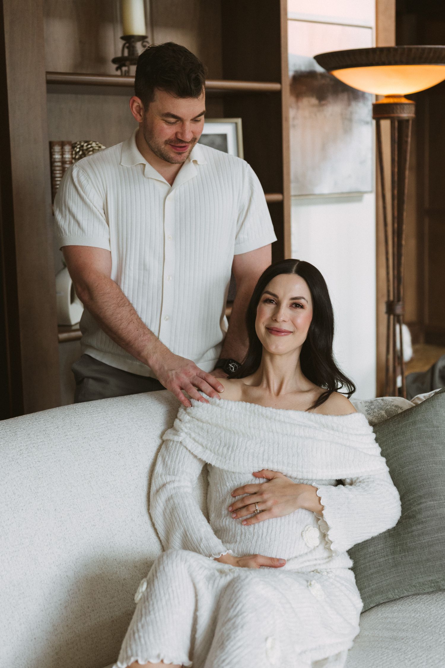 Pregnant woman in white dress sitting on sofa with partner standing behind her indoors.
