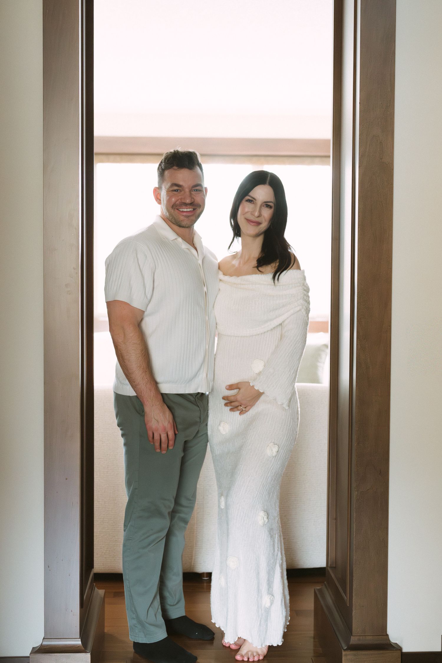 Smiling couple standing in a doorway during a maternity photoshoot, woman in white knit dress.