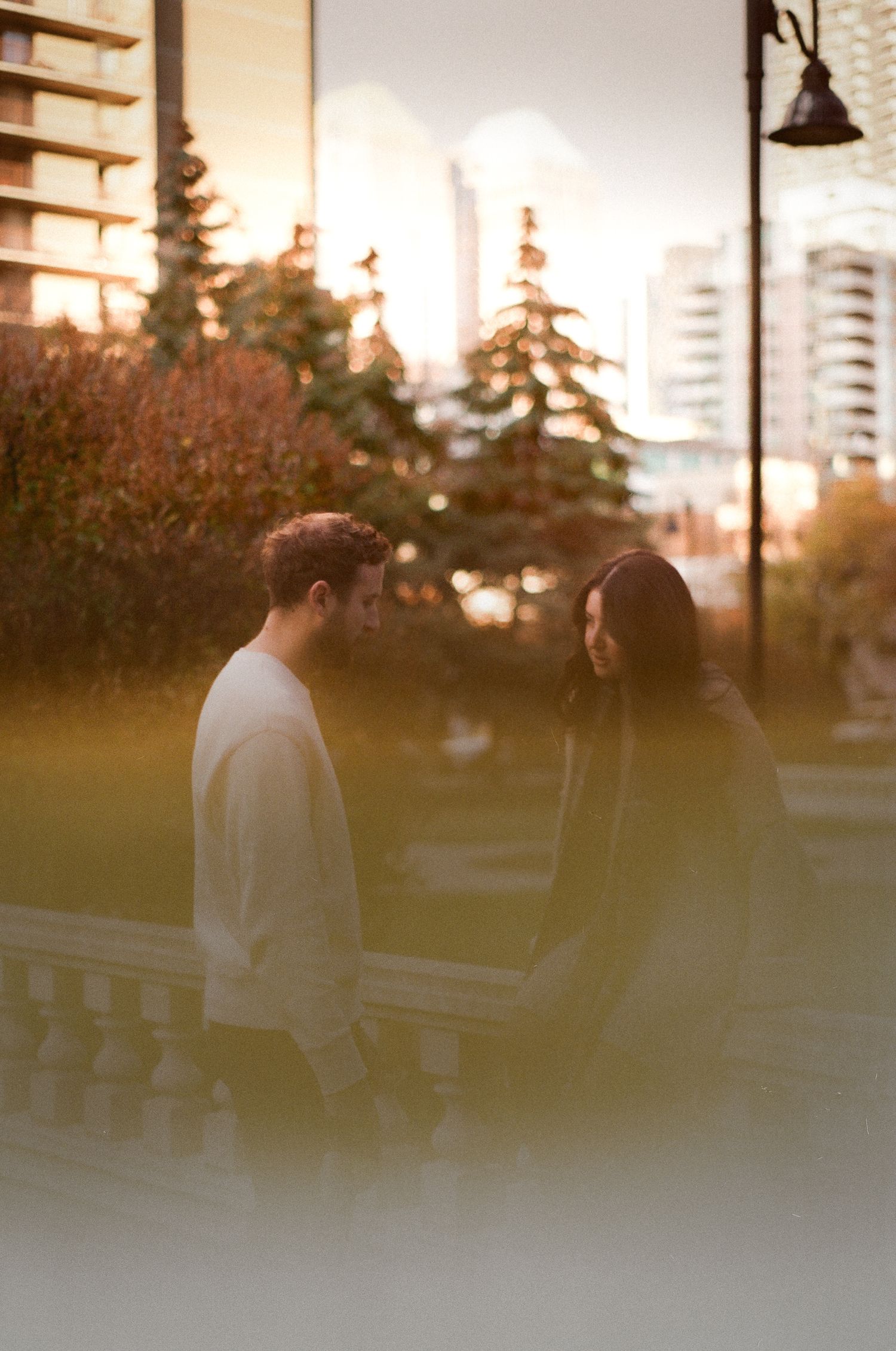 A person in a white sweater walks through a park at sunset with tall buildings visible in the background.