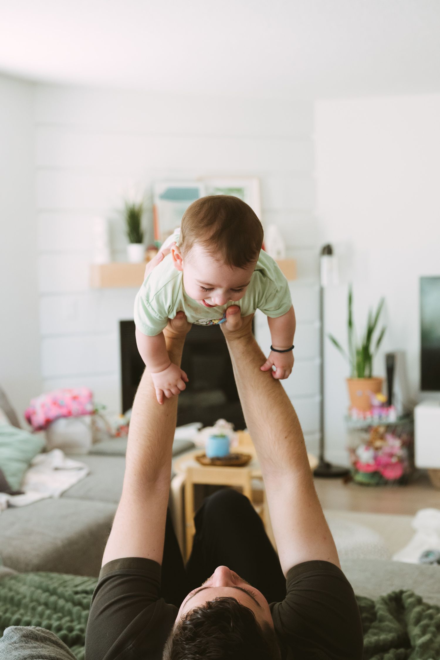A baby is lifted playfully in the air while lying on a couch in a sunlit room with plants.