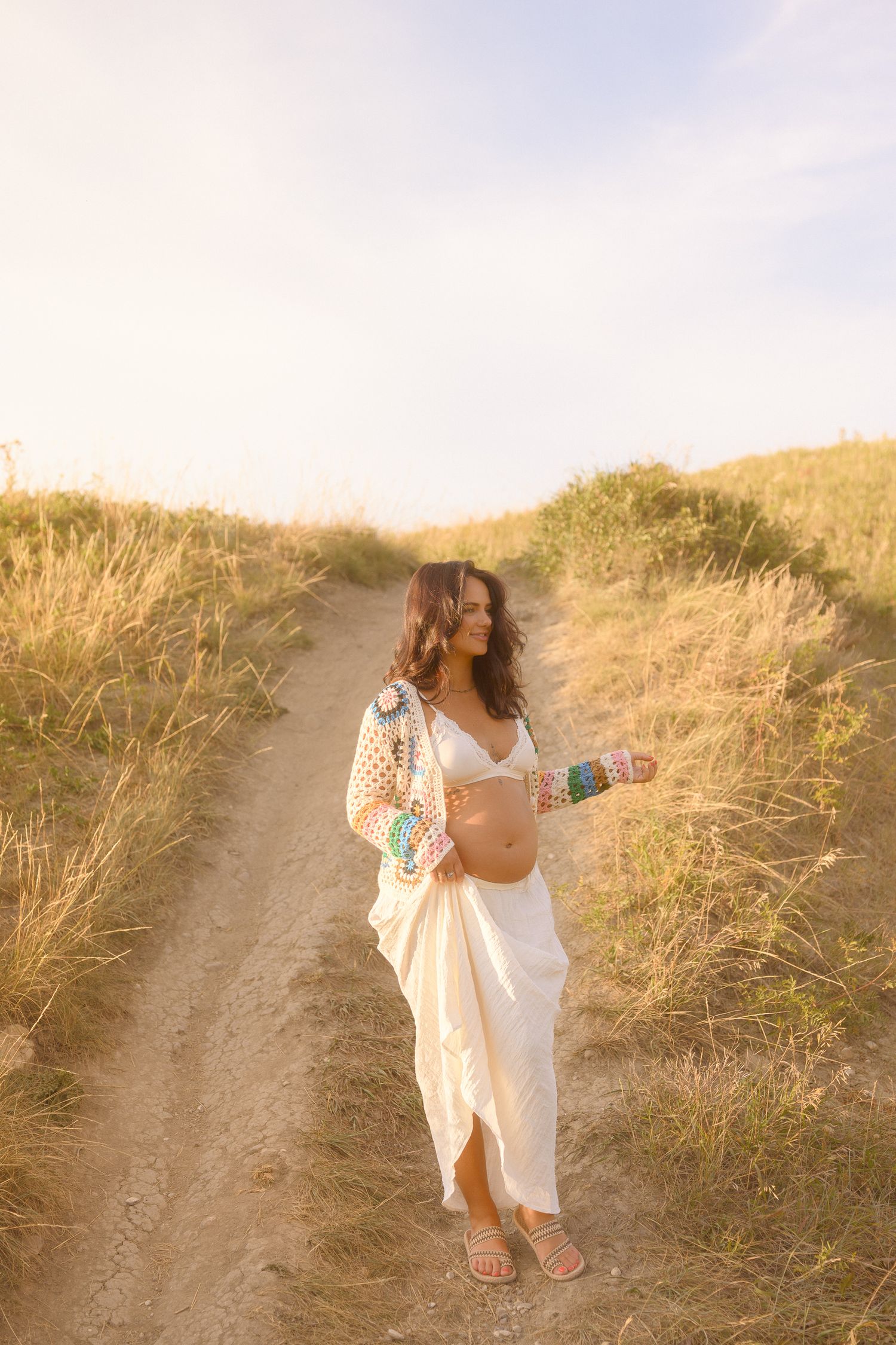 A maternity photoshoot in a golden field shows a flowing white dress against tall grass at sunset.