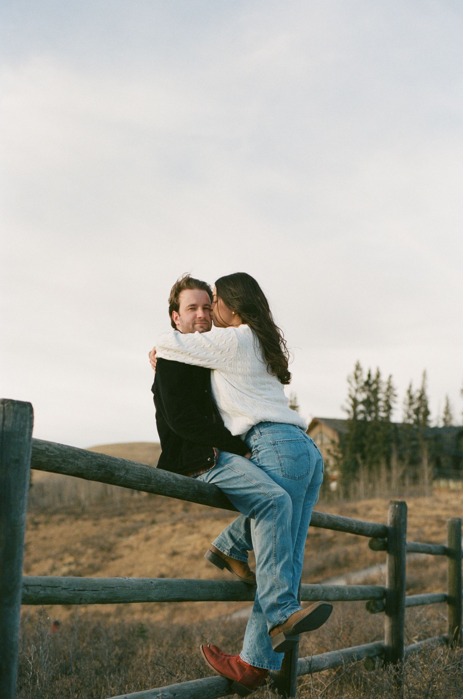 A couple sits together on a rustic wooden fence surrounded by rolling hills and evergreen trees at sunset.