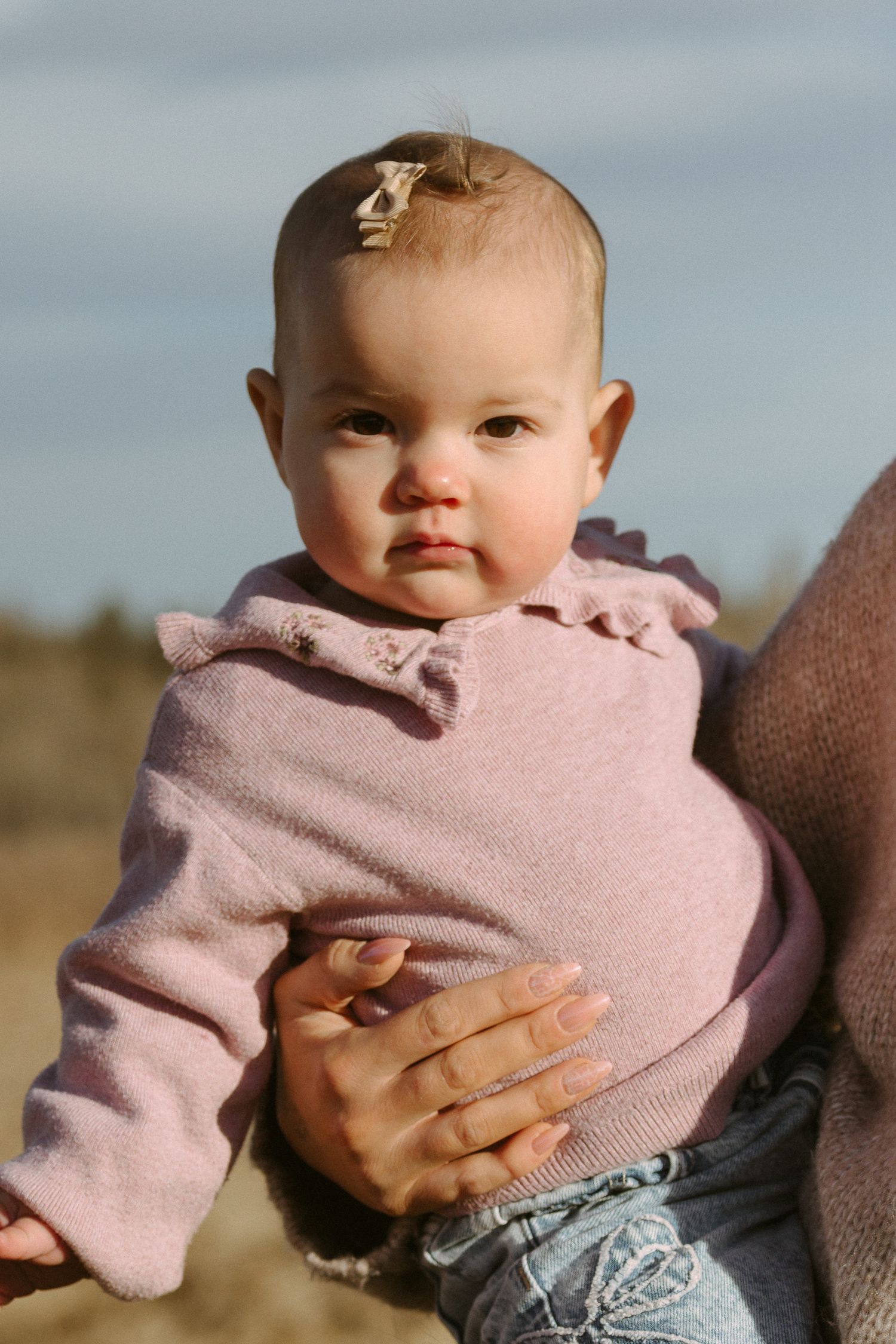 A baby wearing a pink hoodie is held against a cloudy sky background.