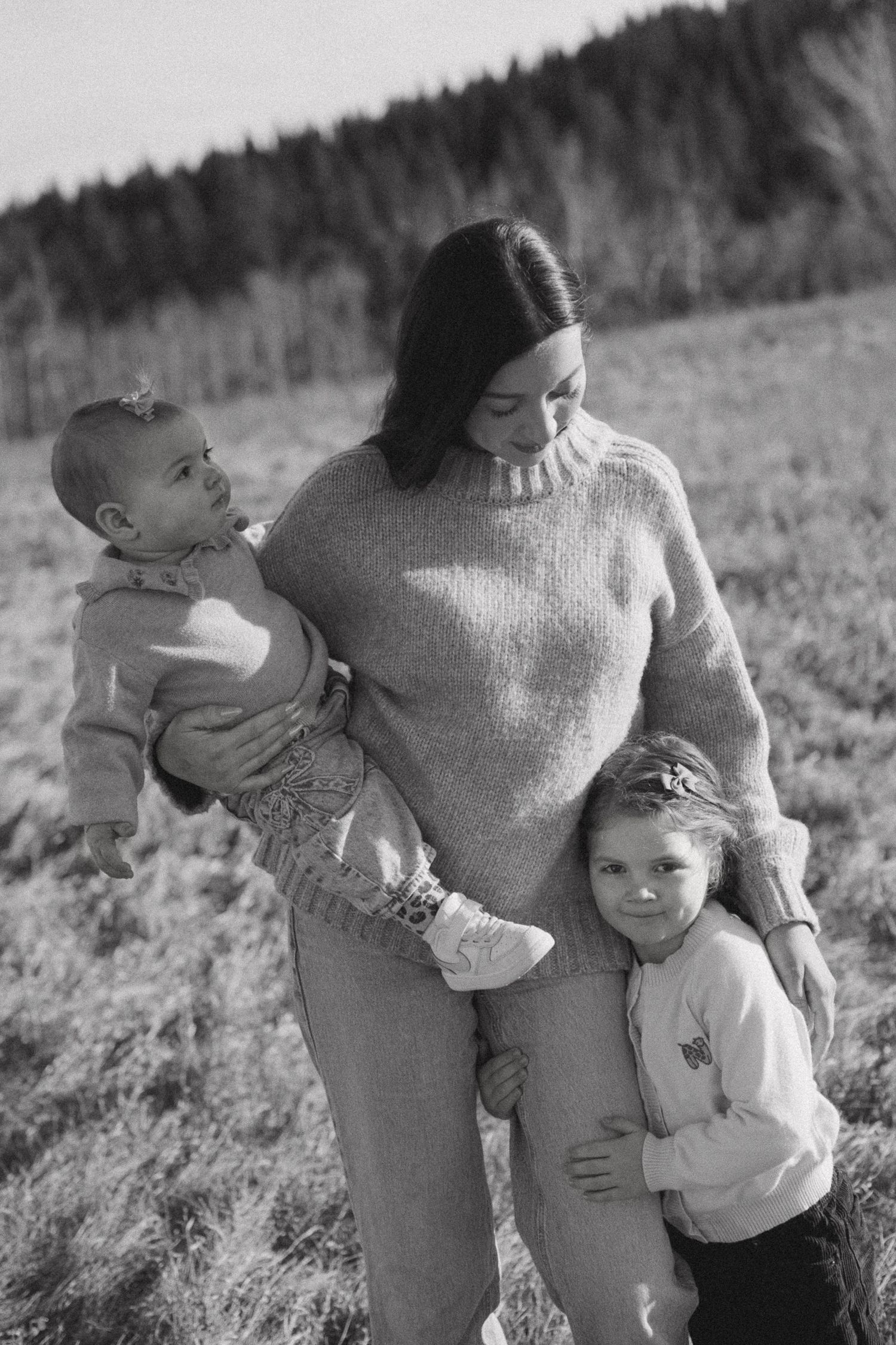A black and white photo of a family embracing in a field with forest backdrop.