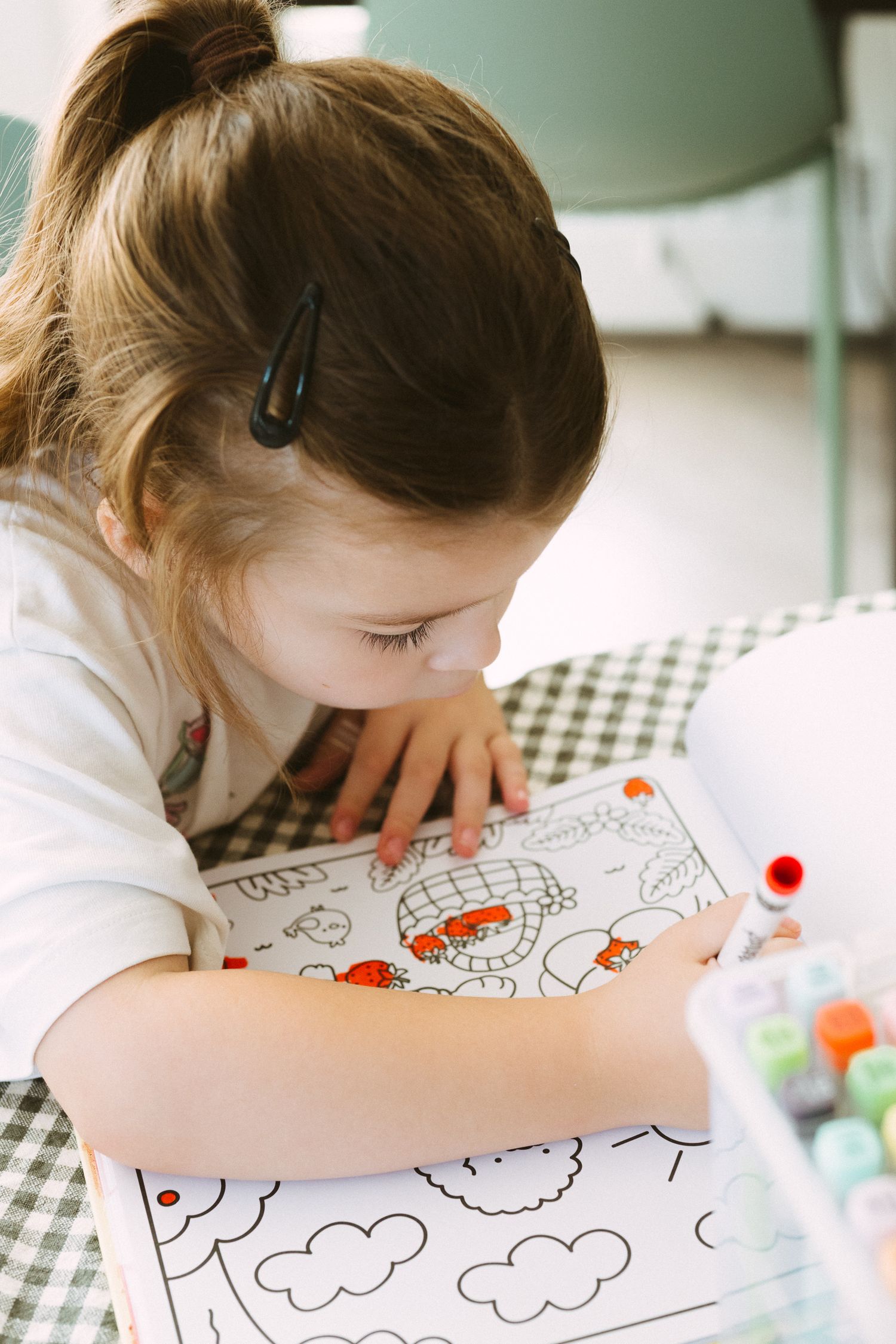 Child leans over paper drawing or coloring with markers while sitting at checkered tablecloth.