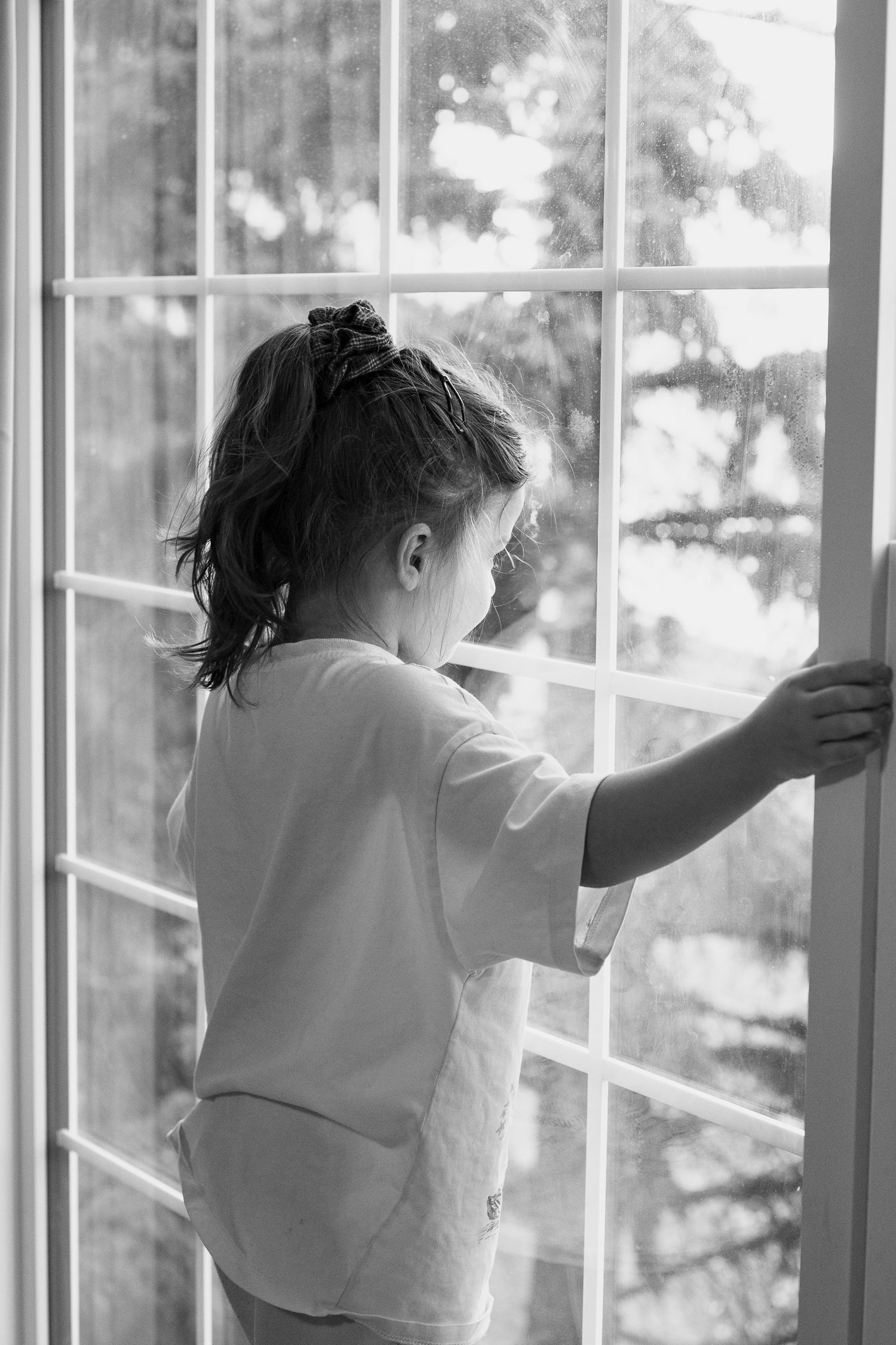 A black and white photo of a small child looking out through a large window with natural light streaming in.