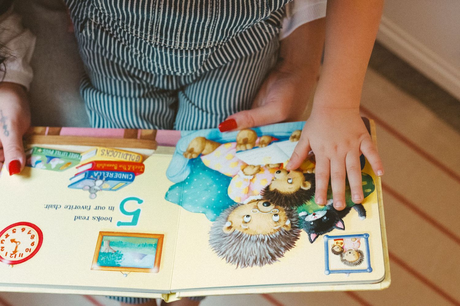 Small hands touch an open children's board book with colorful illustrations on a wooden floor.