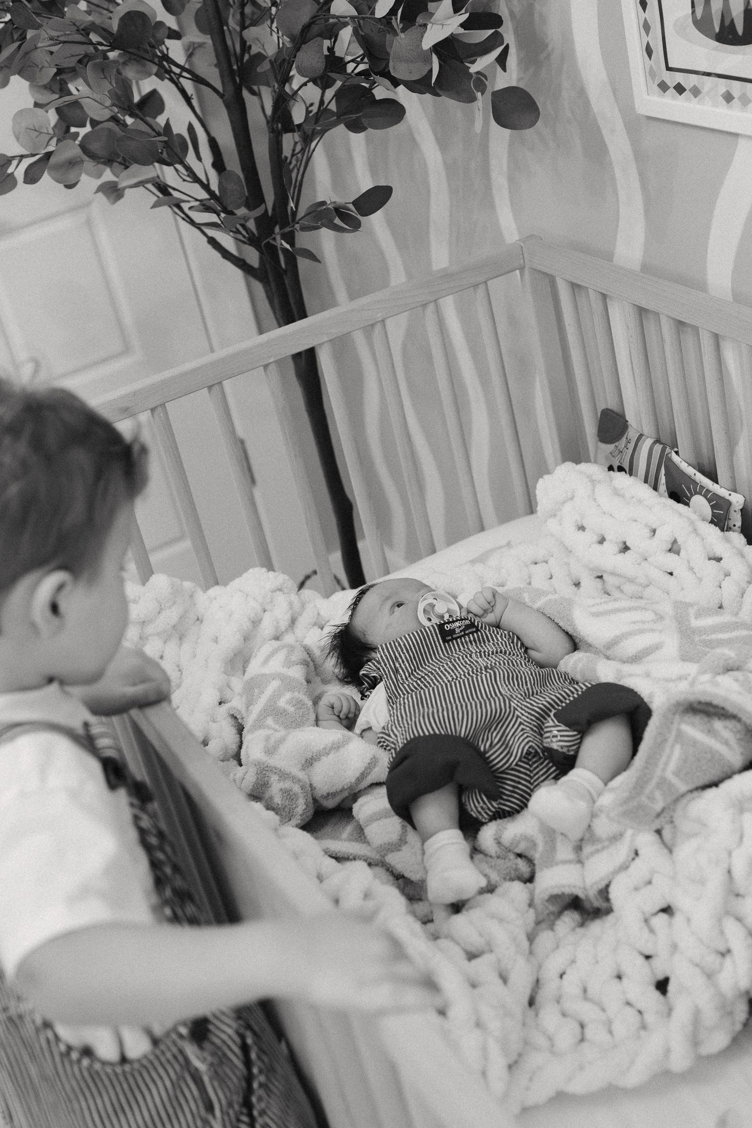 A young child lies in a crib surrounded by plush white bedding and decorative foliage.