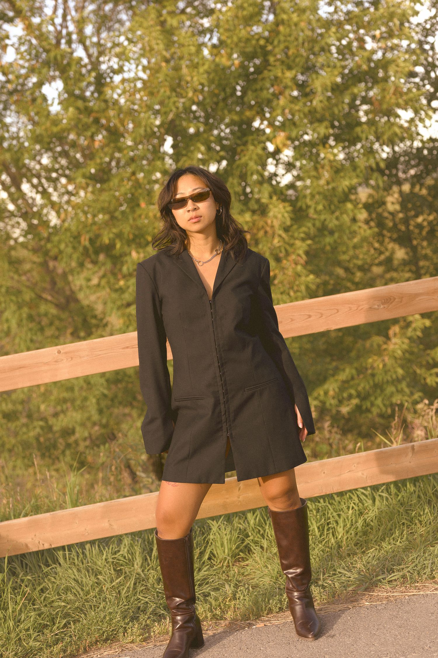 Stylish black blazer dress and boots outfit photographed by wooden fence in natural setting.