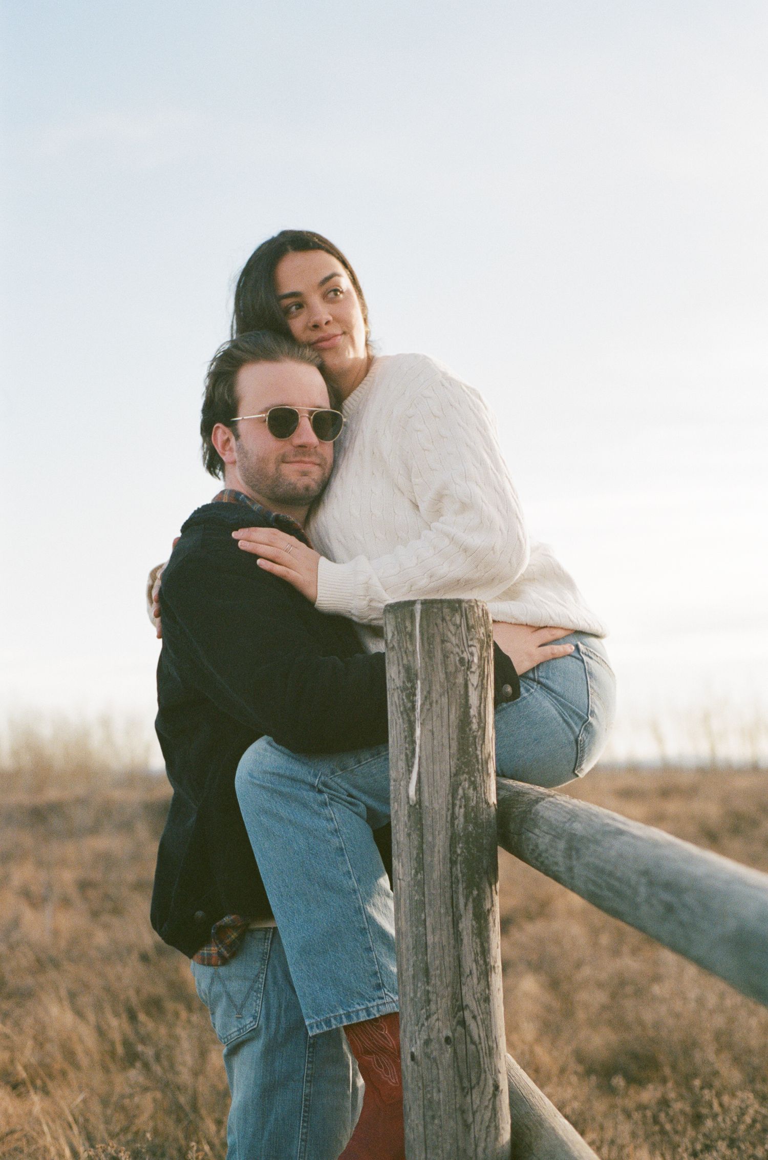 Two people share an intimate moment while leaning against a wooden fence post in nature.