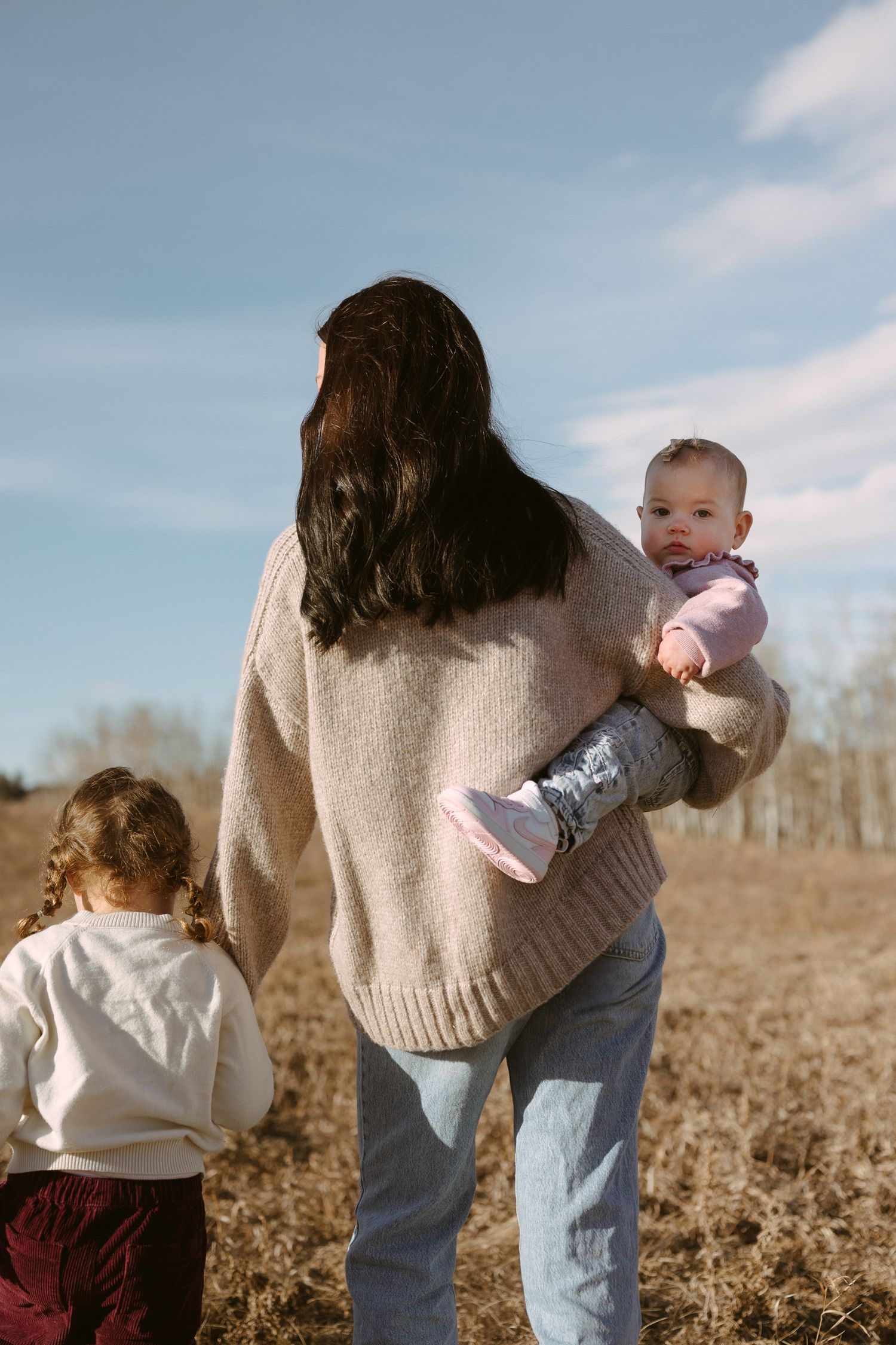 A parent carrying a baby while another child walks alongside in a golden field.
