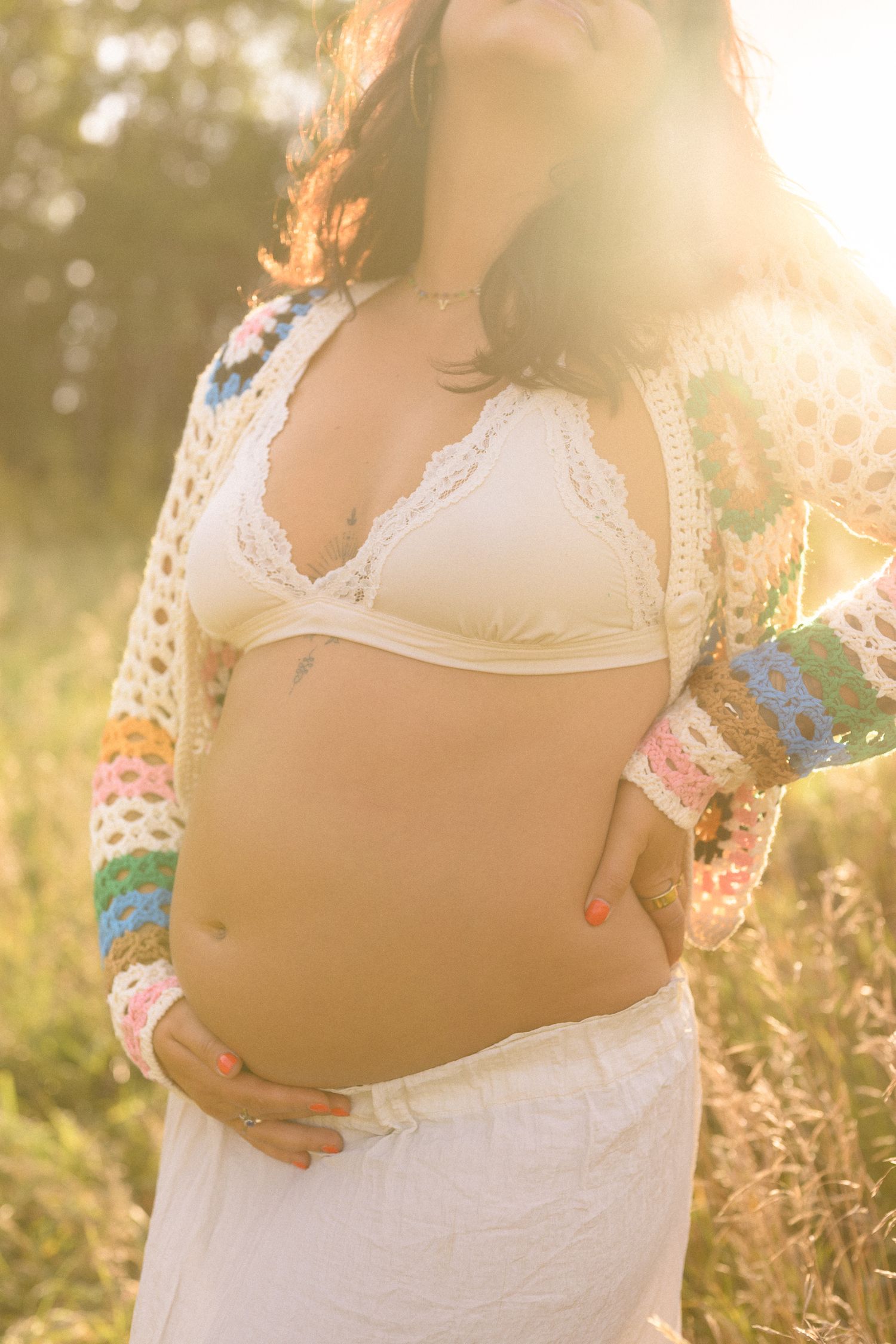 Close-up maternity photos showing a pregnant belly in white lace against sunlit meadow flowers.