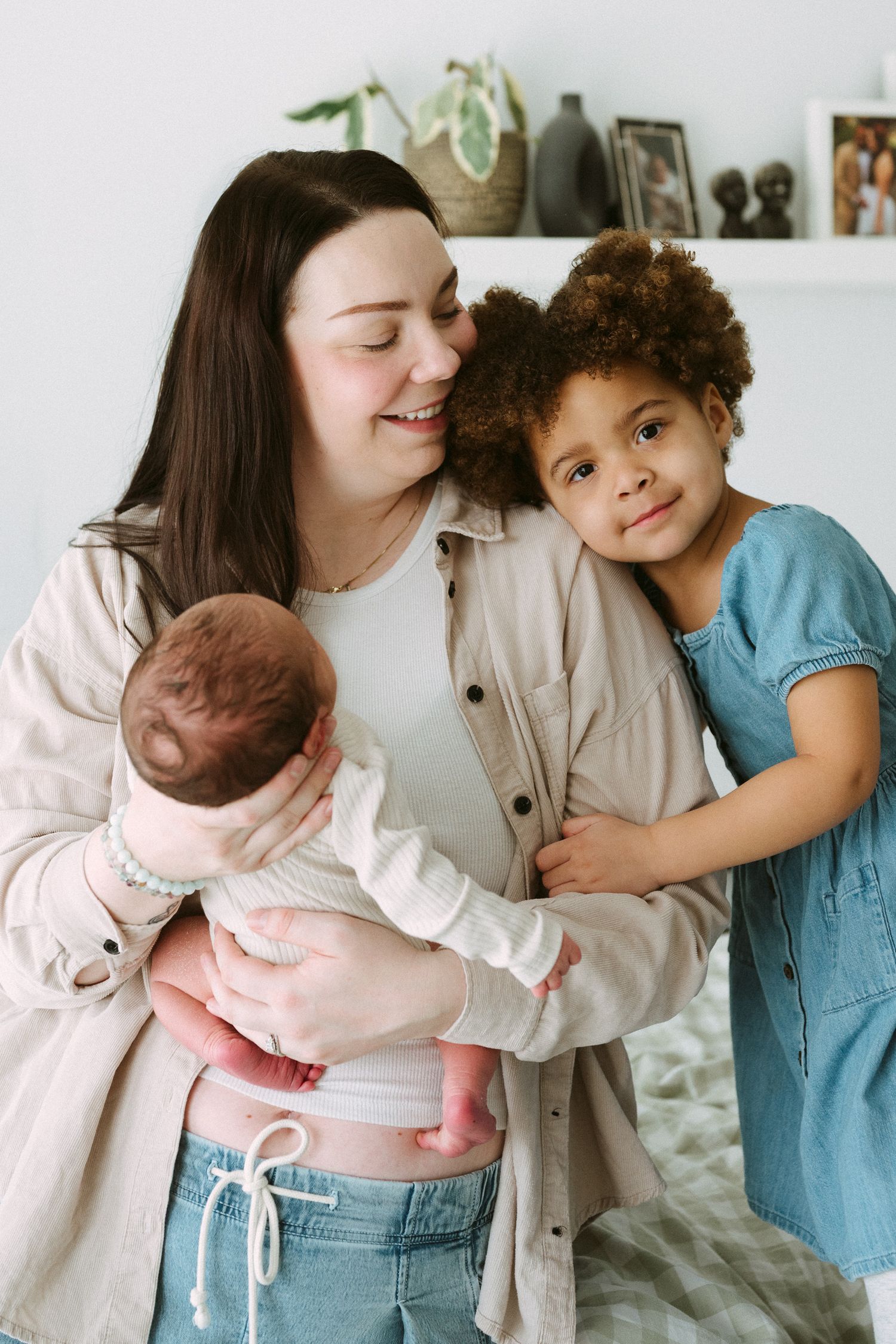 A smiling mother holds her newborn baby while an older curly-haired girl in a blue dress hugs her warmly indoors.